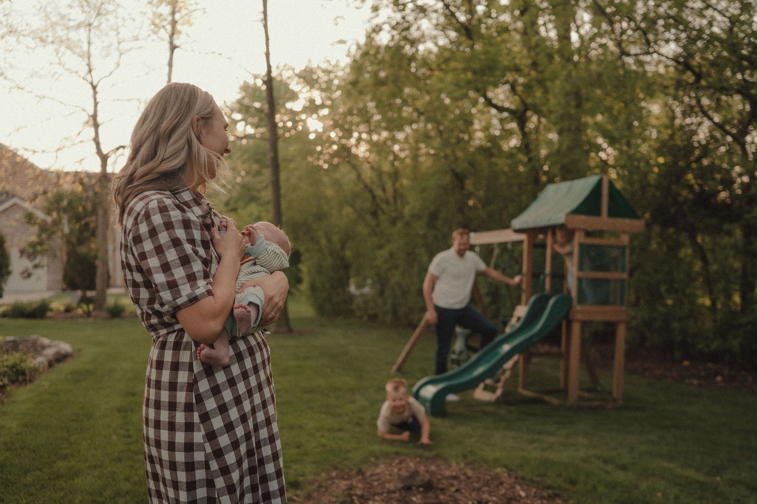 A mother holds her newborn in a plaid dress while her family plays near a backyard swing set.