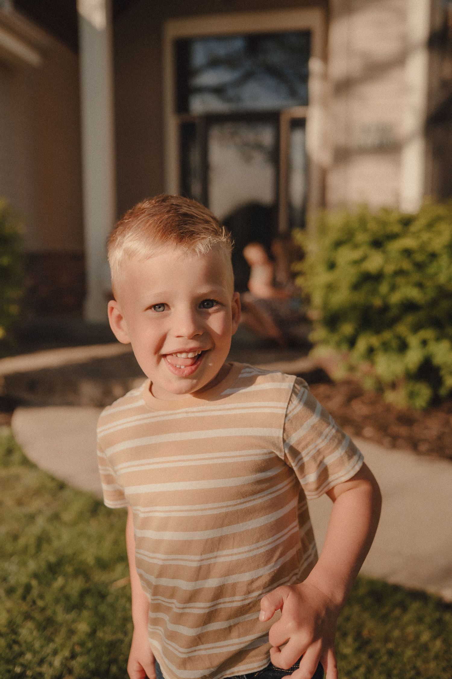 Happy blond toddler boy smiling in striped shirt on green lawn outside home.