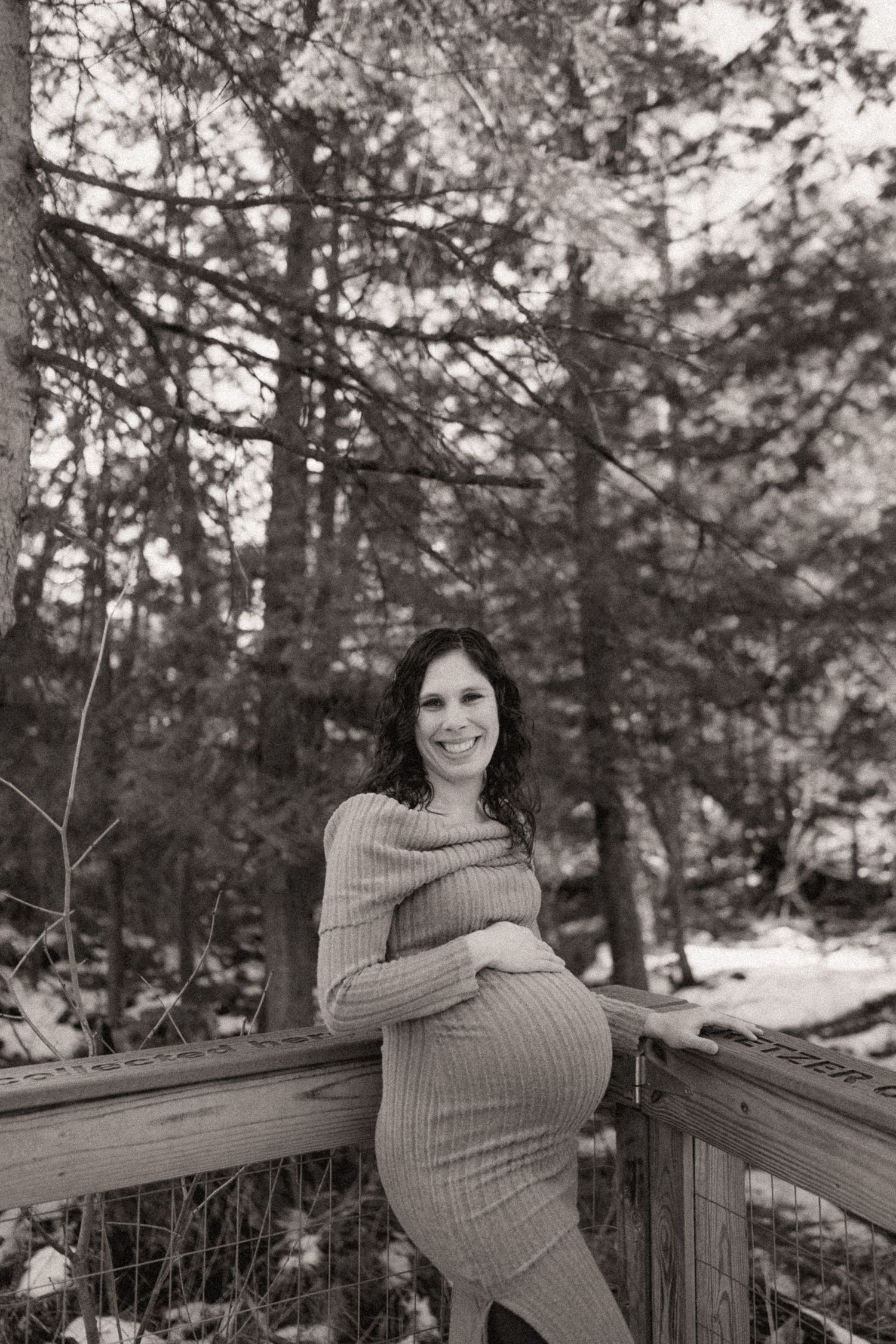 A black and white portrait of someone in a knit dress sitting on a wooden railing in a forest setting.
