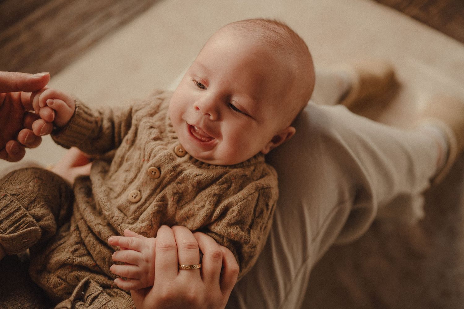 A baby wearing a cozy beige knit sweater smiles while lying down on white bedding.