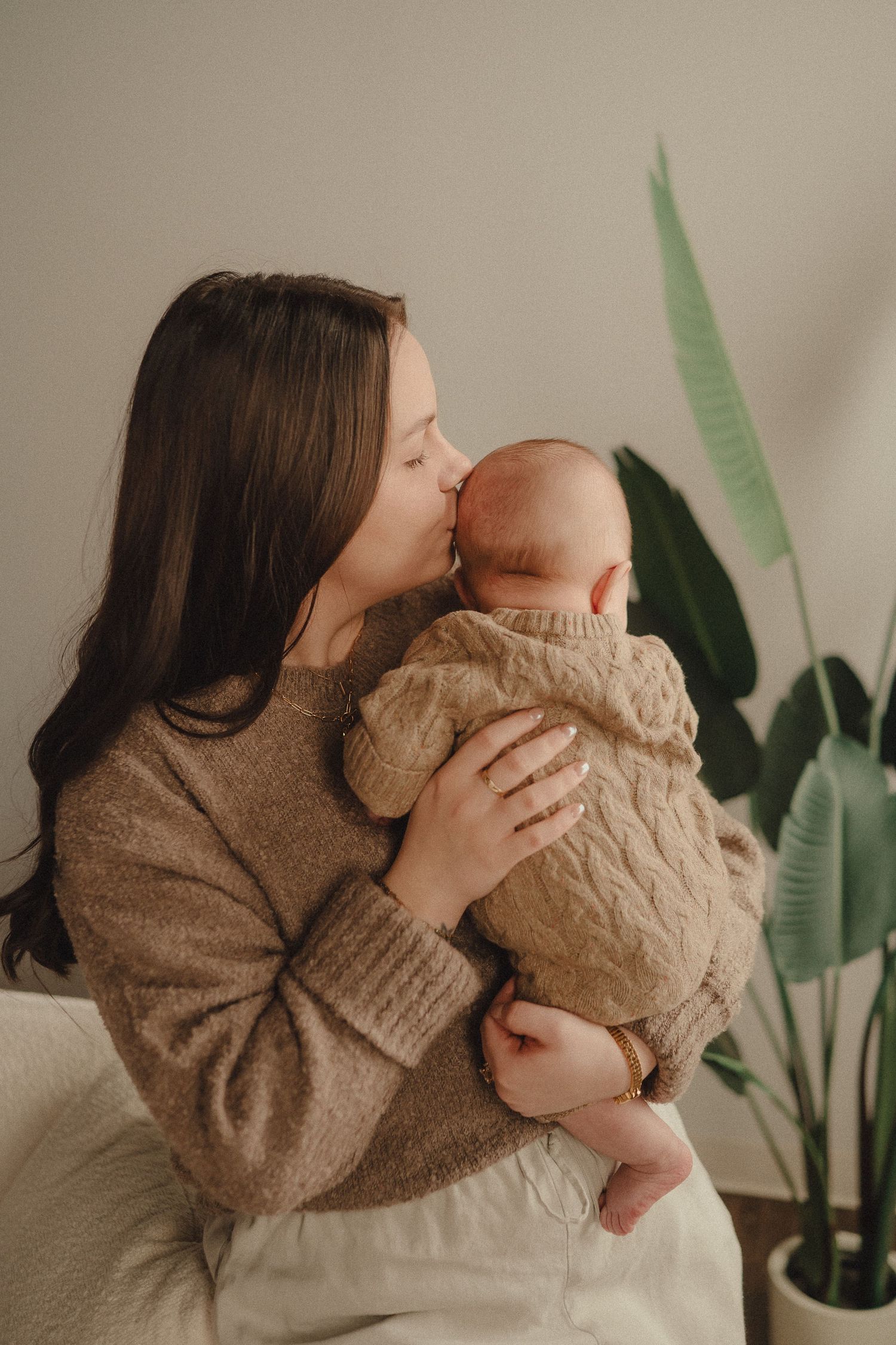 A series of tender moments captured in warm tones showing parent and baby in matching knit sweaters near a houseplant.