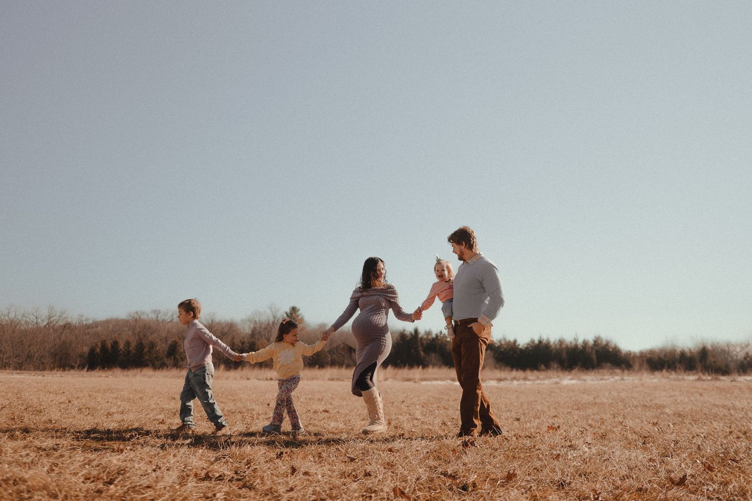 Family holding hands and dancing together in a golden field during sunset on a clear day.