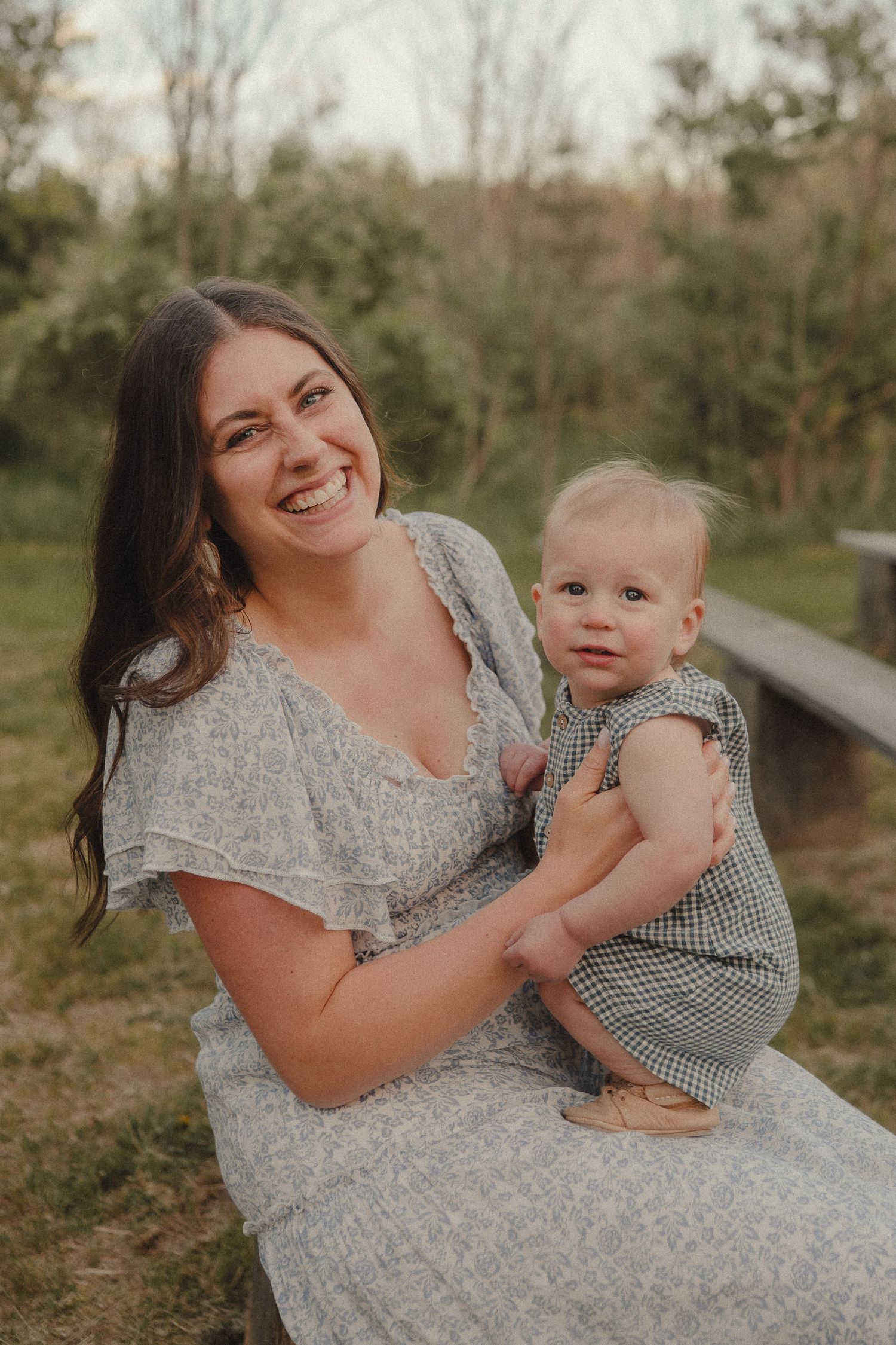 Smiling brunette mother in grey dress holds her baby girl on a wooden bench outdoors at golden hour.
