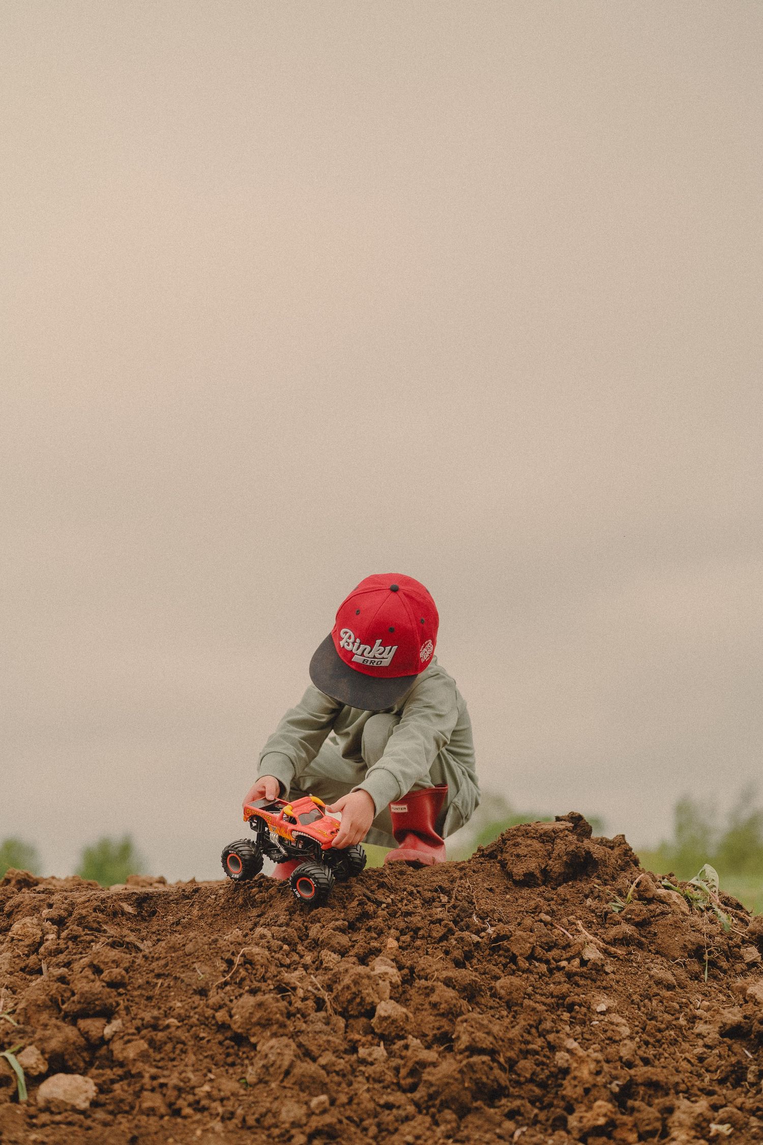 Young boy in red cap playing with orange monster truck toy in dirt outdoors on overcast day.