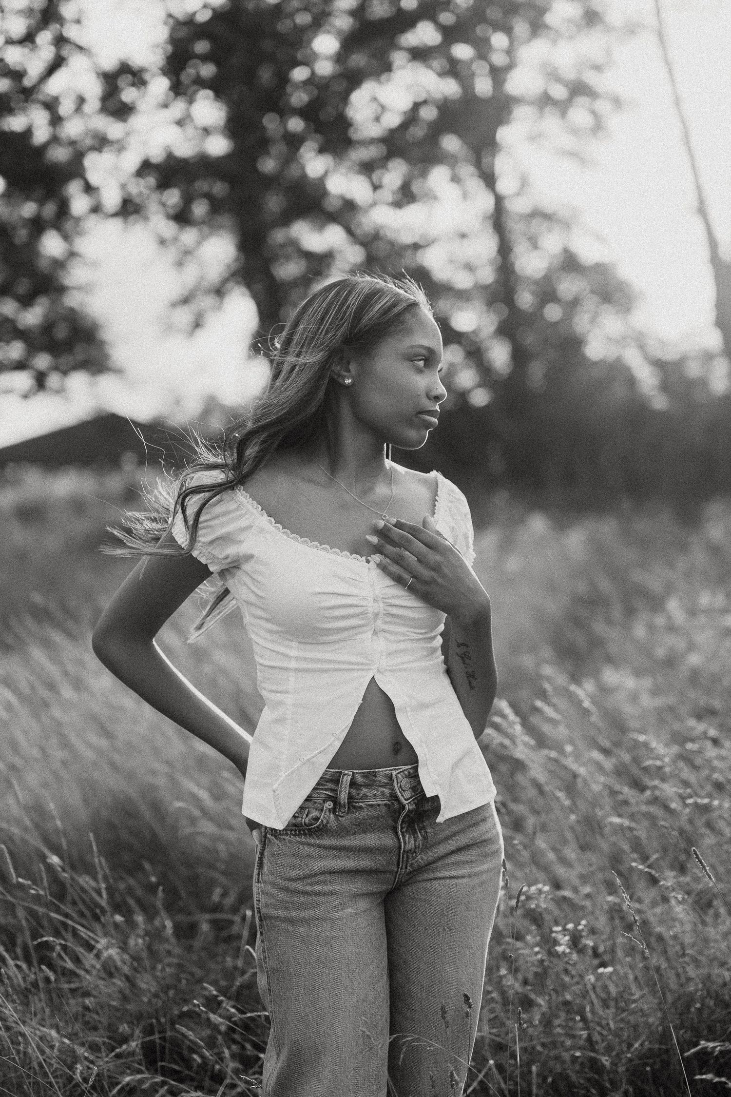 Black and white photo of a young woman in a white crop top and jeans standing in a field with trees in background.