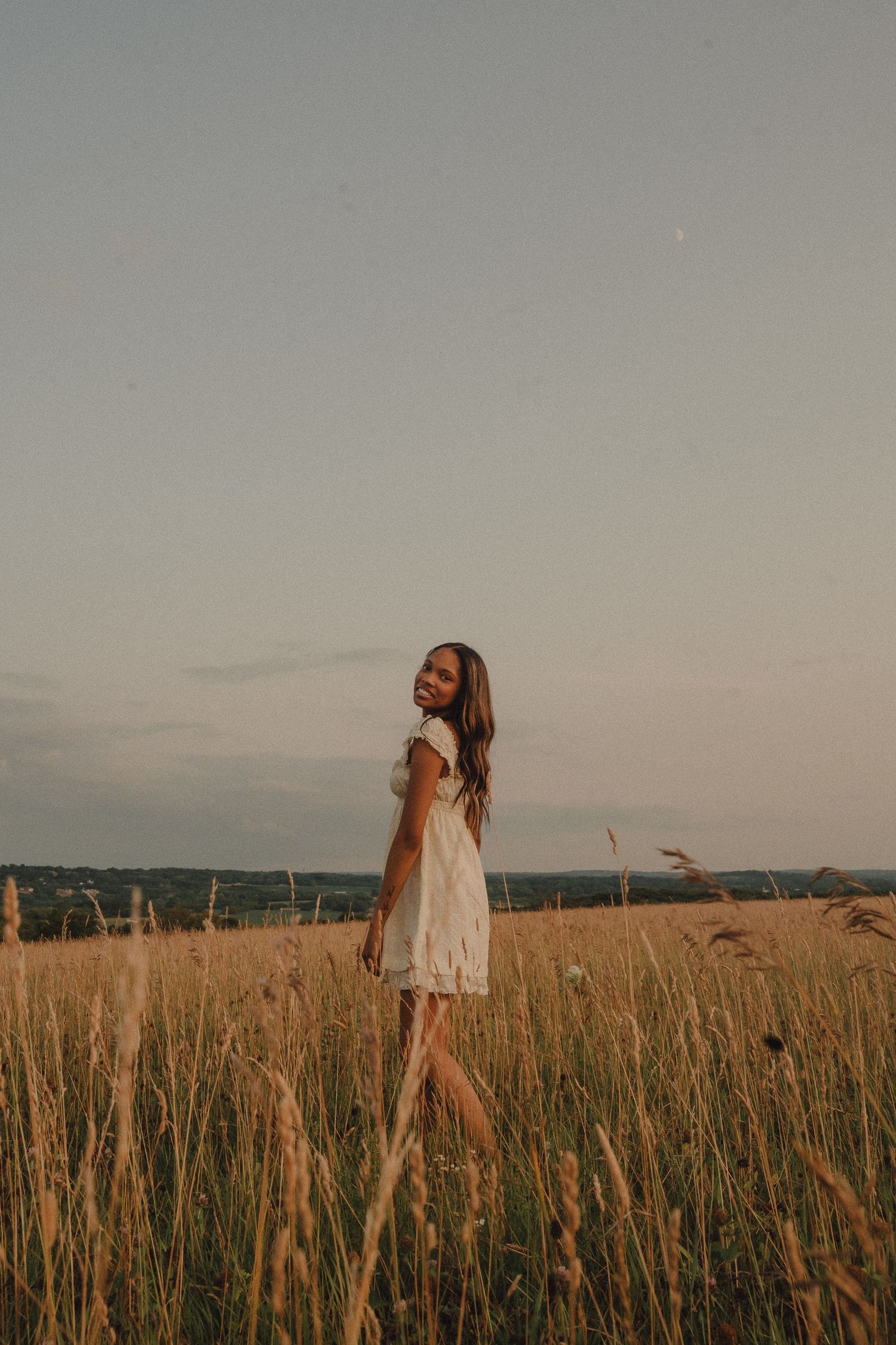 Young woman in white lace dress smiling over shoulder in golden wheat field at dusk.
