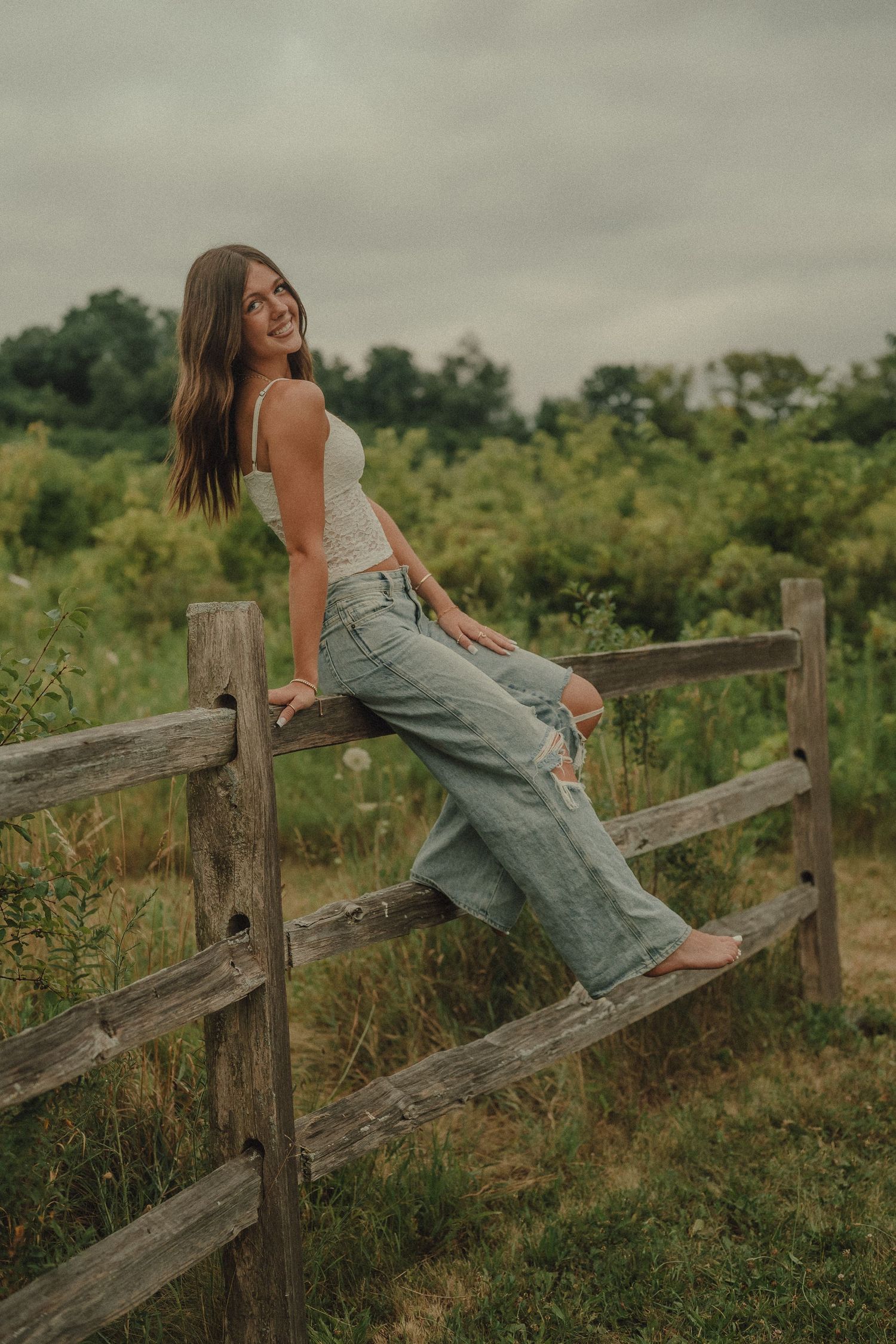 Smiling young woman in white tank and ripped jeans sitting on rustic wooden fence in countryside.