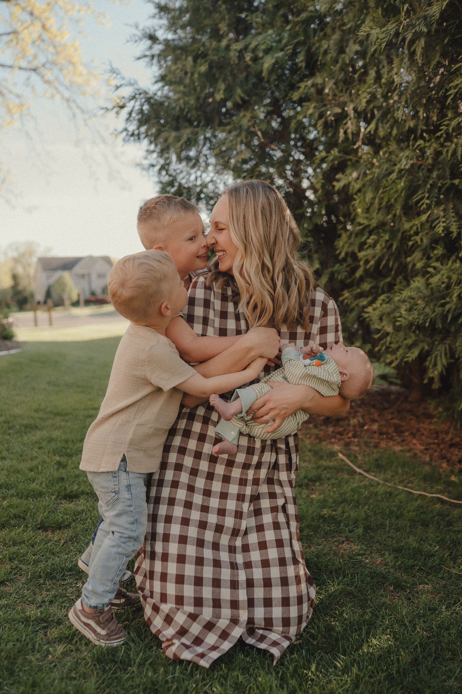 A smiling mother hugs her two young sons while holding a newborn baby in a sunny backyard.