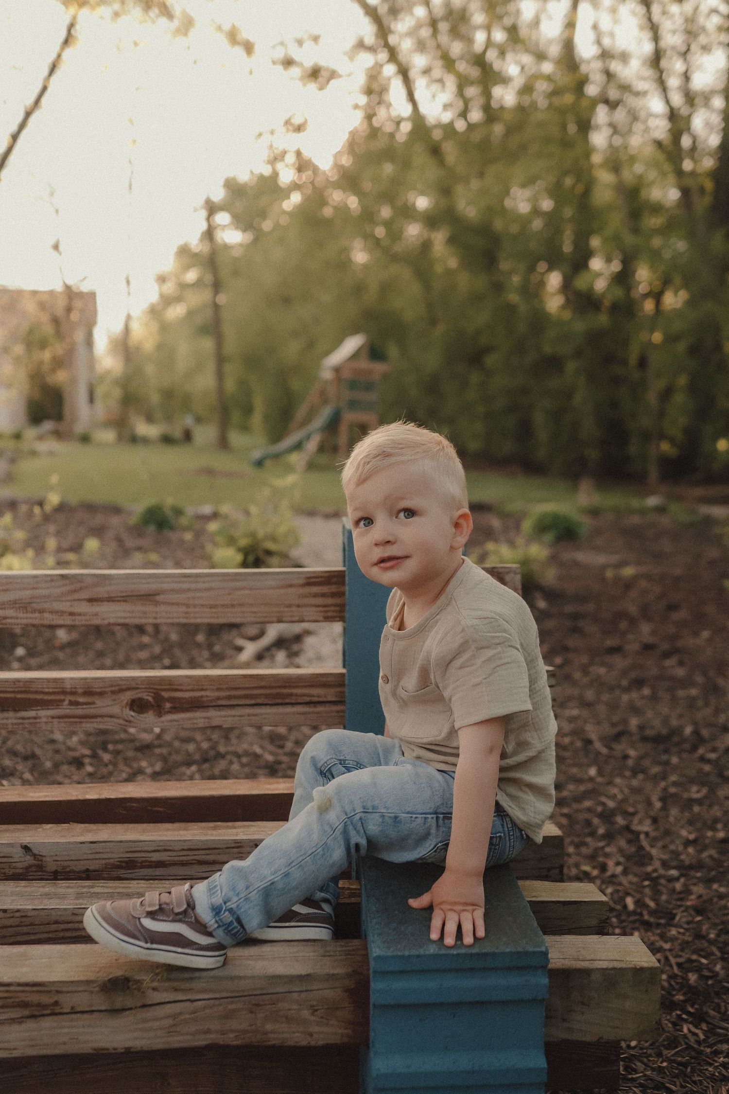 A young blonde toddler boy in jeans sits on wooden garden steps in warm afternoon sunlight.