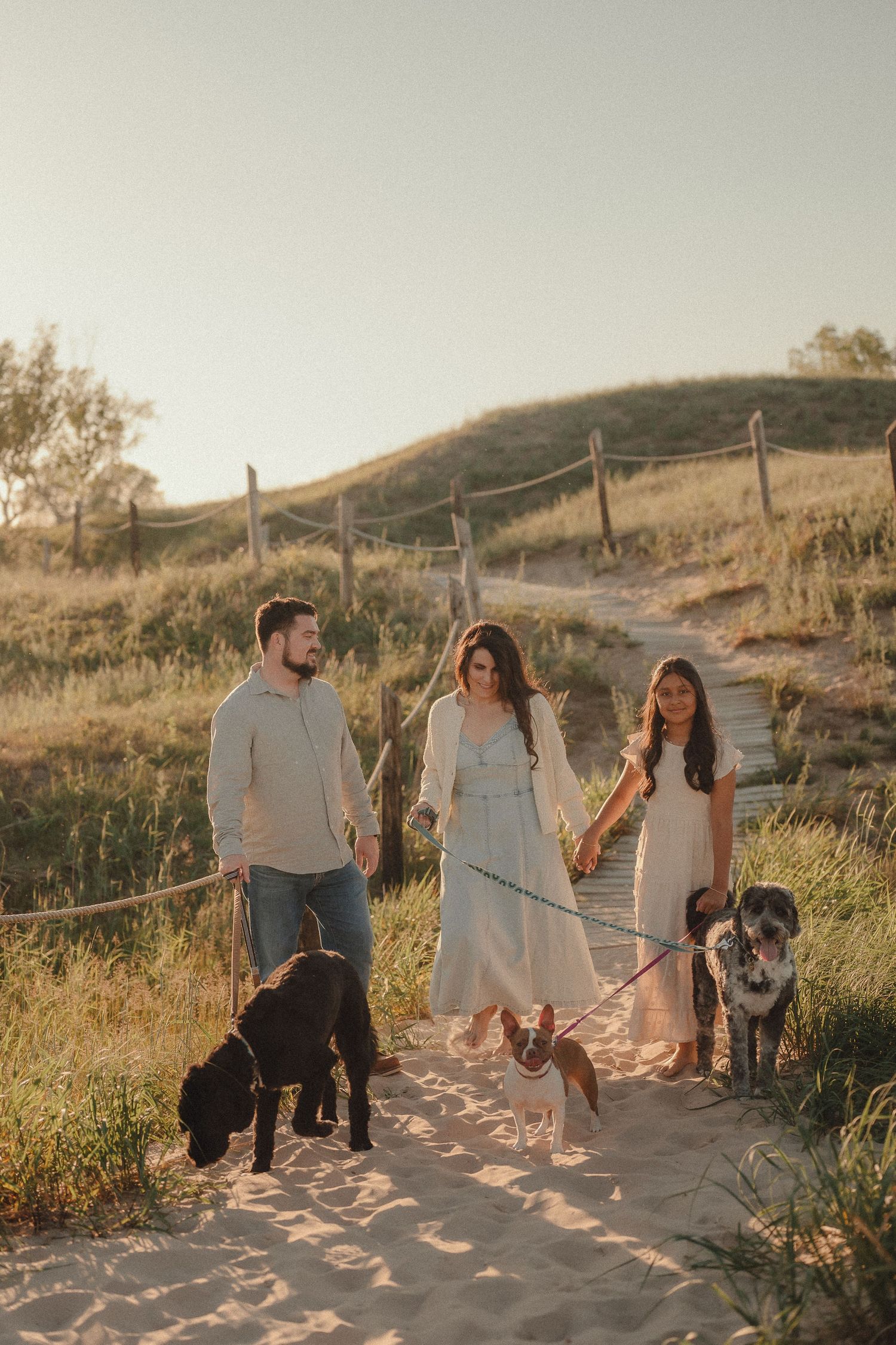 Family walking dogs on sandy beach path at golden hour with green hills and rustic fence in background.