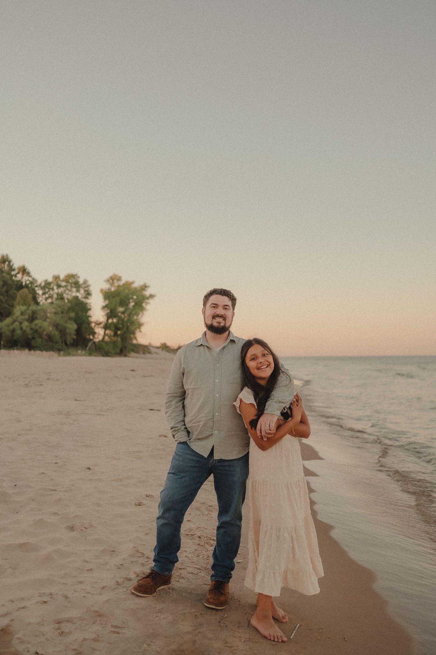 Couple posing together on sandy beach at sunset, woman in white dress leaning on mans shoulder.