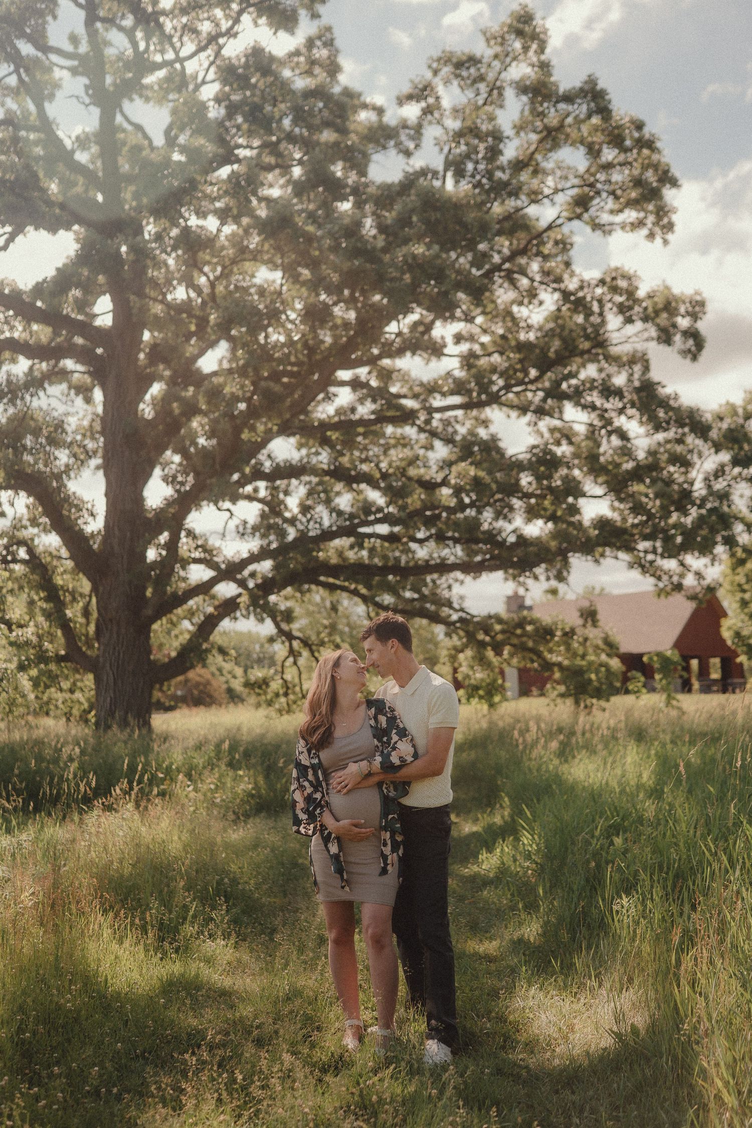 A couple embraces tenderly in a sunlit meadow with a large oak tree and red barn.