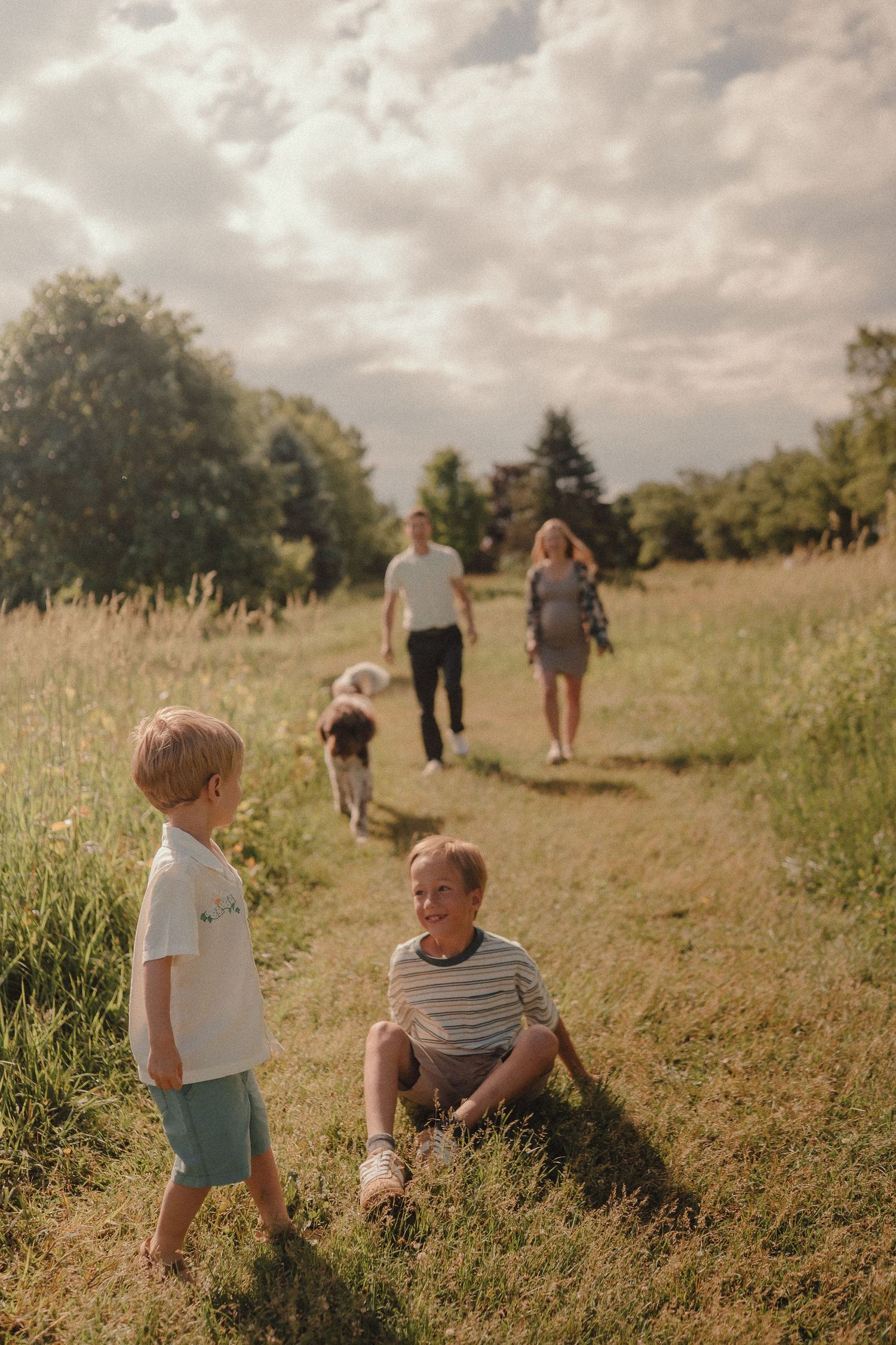 Family with two boys and a dog walking through a sunny green meadow under a cloudy sky.
