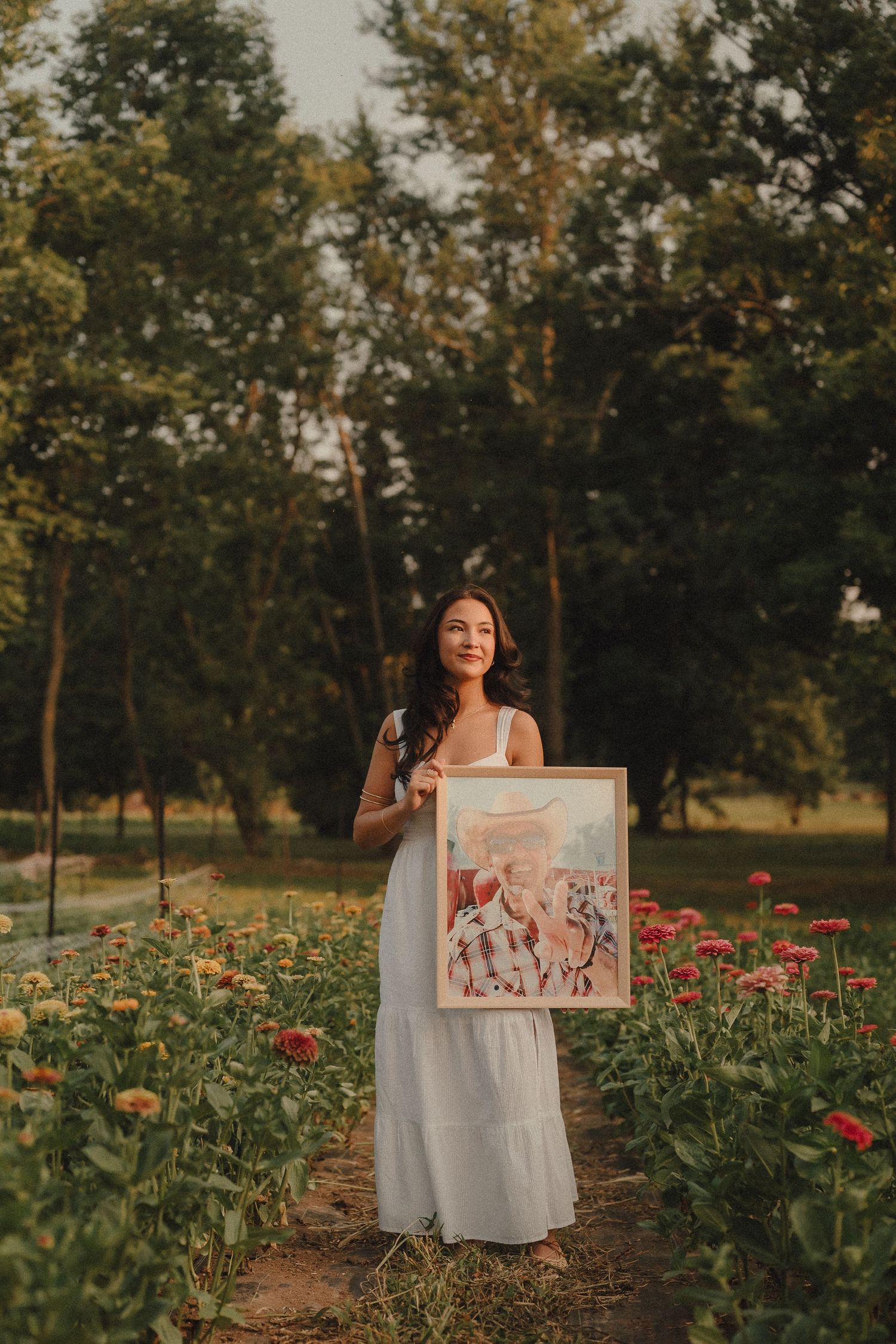 Woman in white dress holding a framed painting in a colorful flower garden with lush green trees.