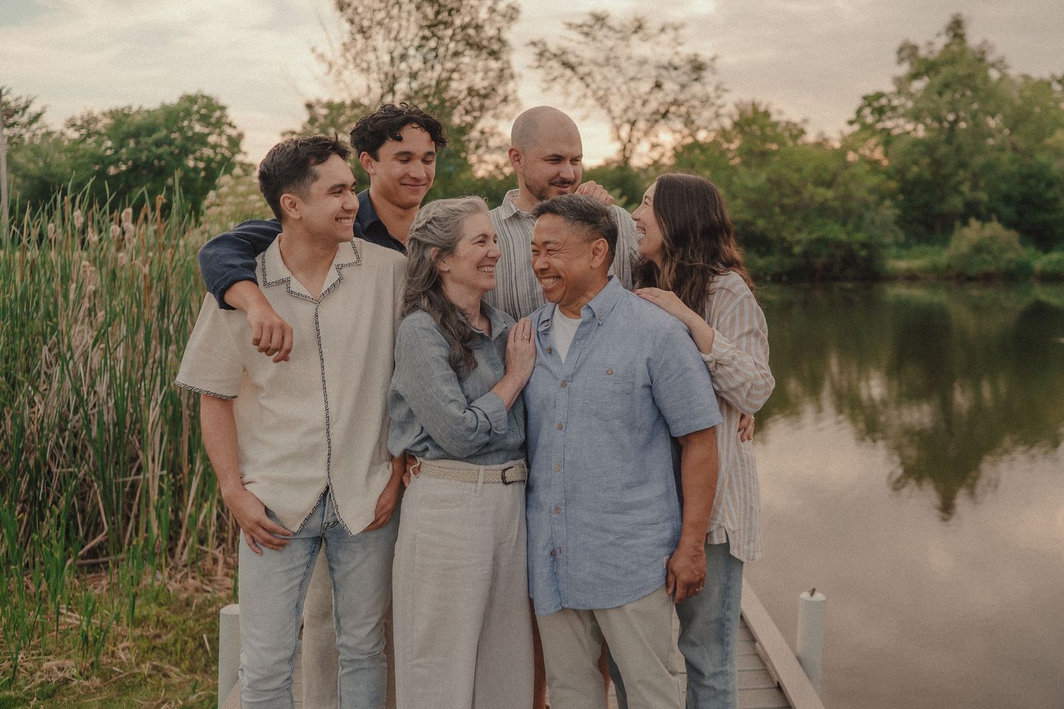 Happy multigenerational family laughing together outdoors by a serene lake surrounded by lush green trees at sunset.