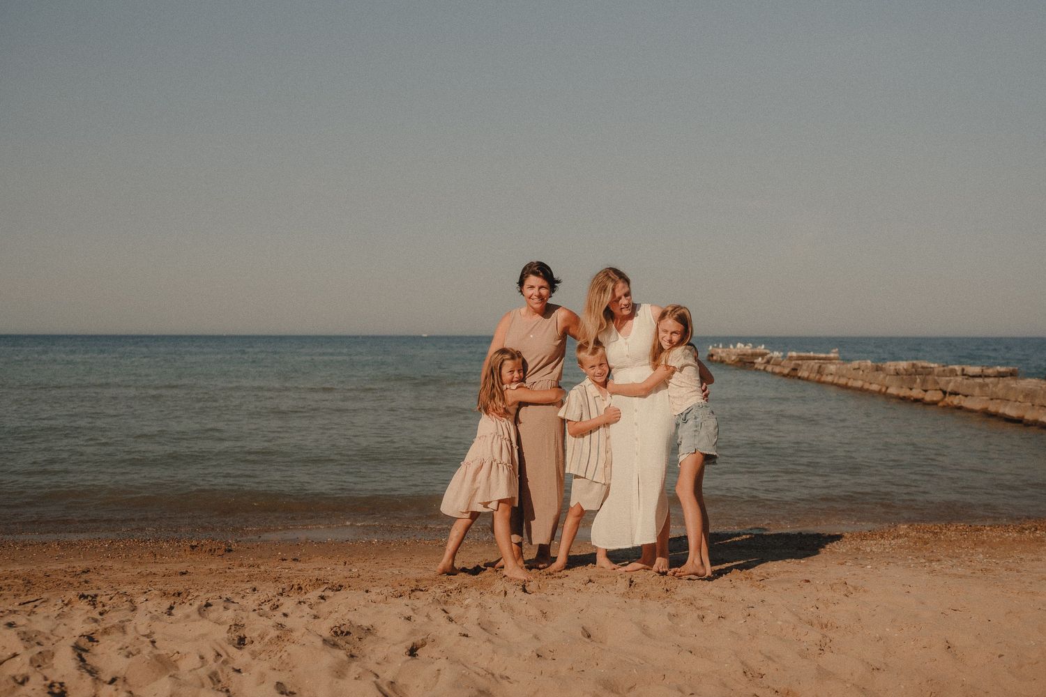 A joyful family of four in light-colored dresses embraces together on a sandy beach by the ocean.