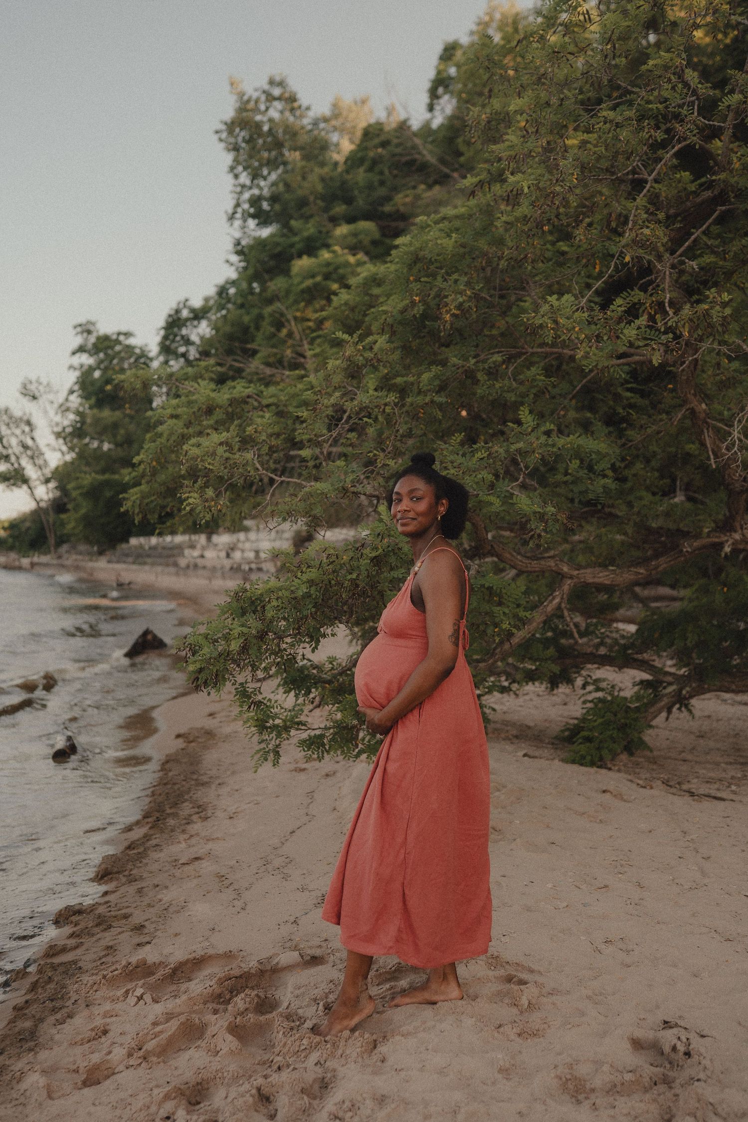 Smiling pregnant woman in coral dress standing barefoot on sandy beach near lush trees.