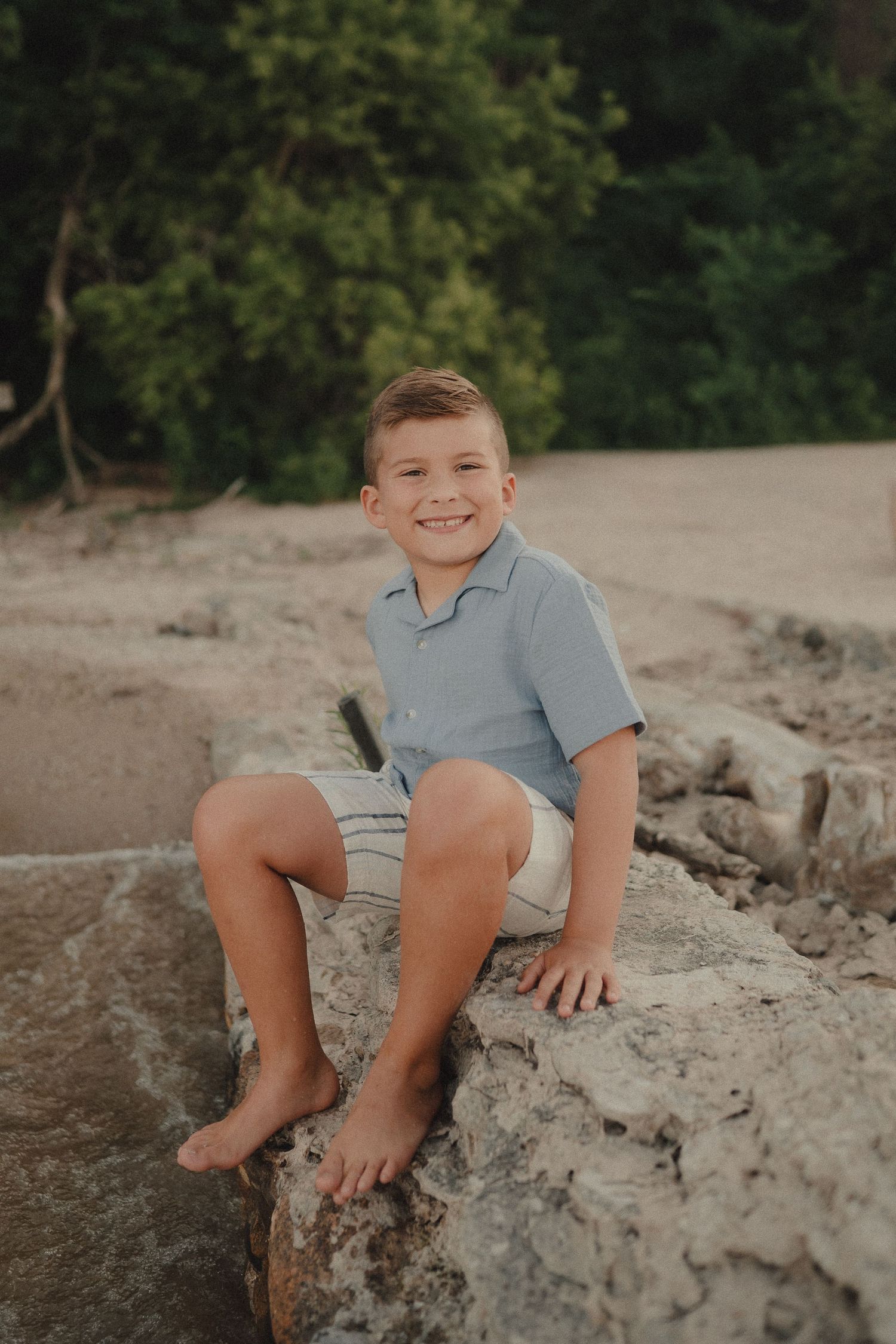 Smiling boy sitting on rocks at the beach wearing a light blue shirt and striped shorts.