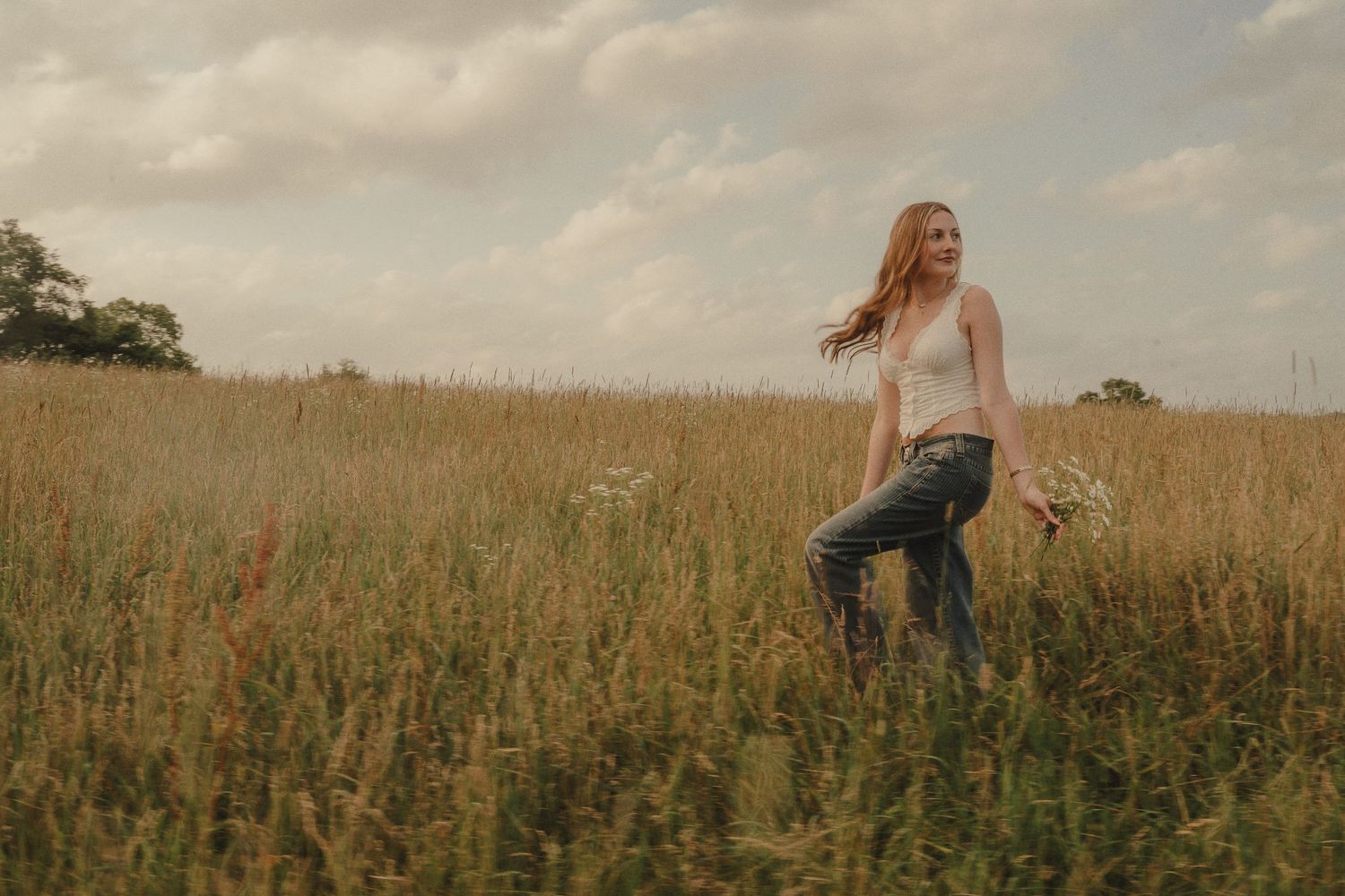 Young woman in jeans and white top walks through tall golden field holding wildflowers on cloudy day.