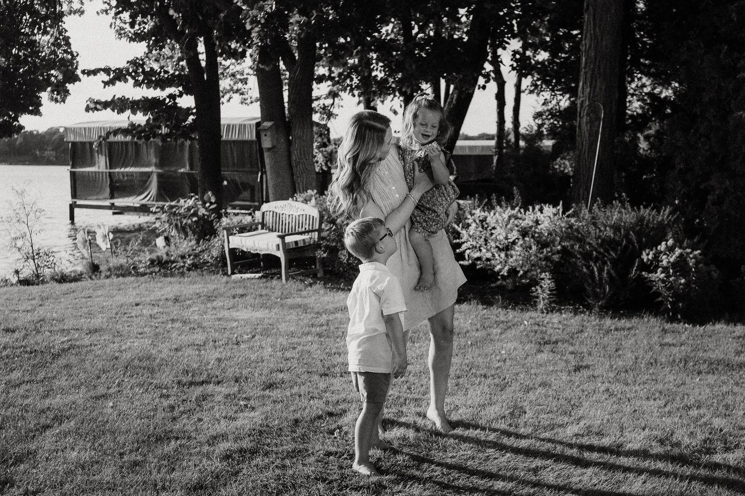 Black and white photo of a woman holding two young children in a lush backyard near a lake.