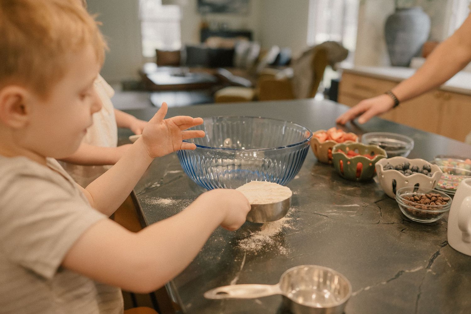 Young child helping bake in kitchen, mixing ingredients in blue bowl on dark countertop with measuring cups nearby.