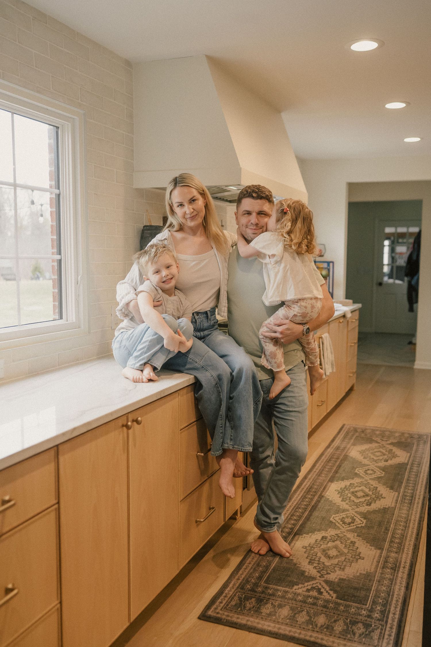 A happy family of four poses in a modern kitchen, with parents holding two young children near a marble countertop.
