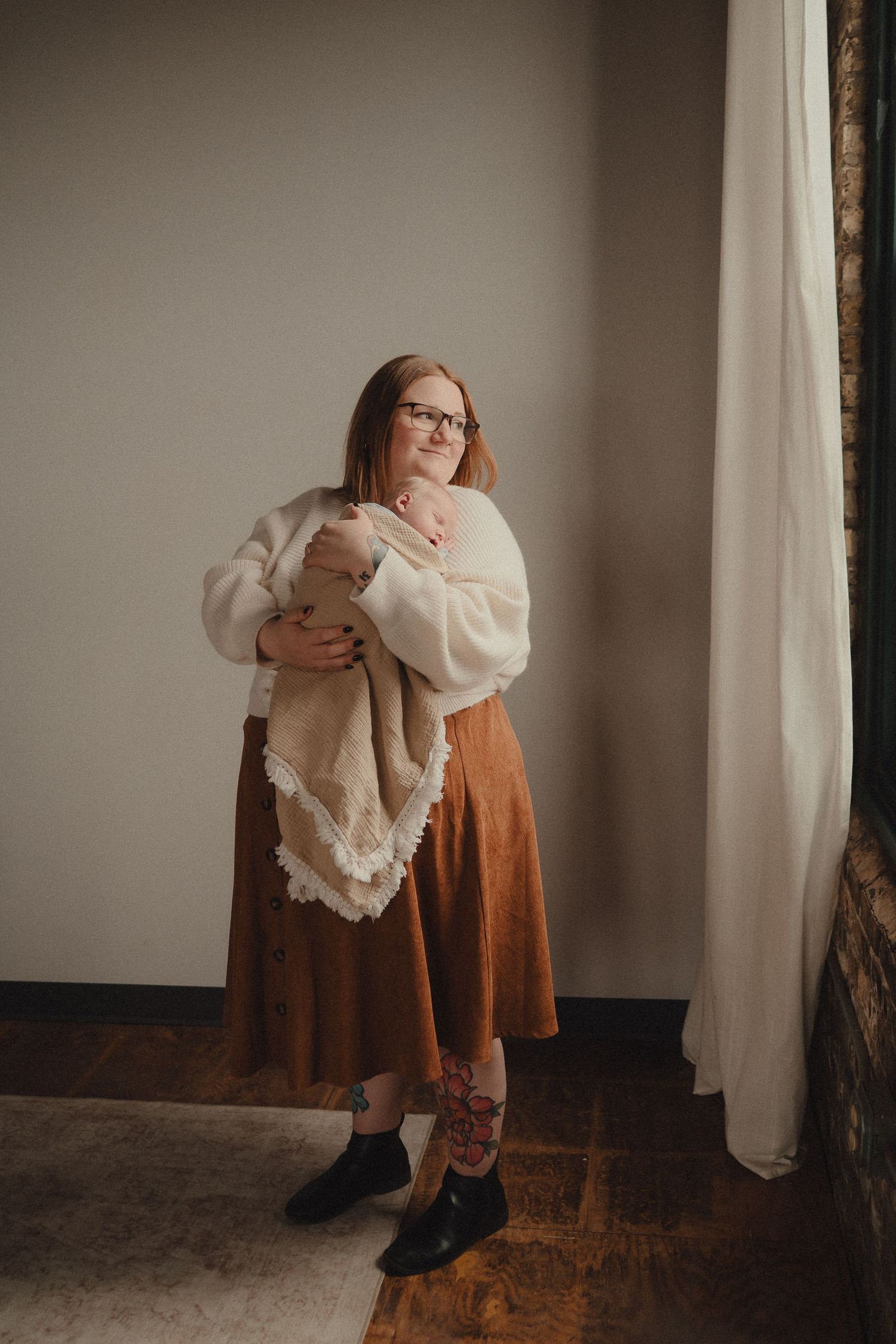 Mother wearing glasses and rust skirt holds swaddled newborn baby near a bright window indoors.
