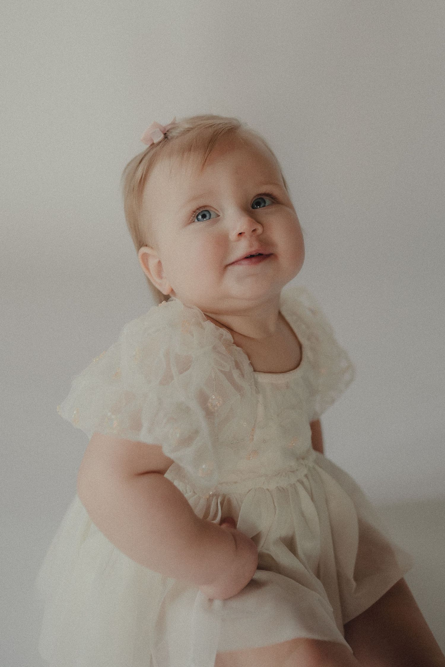 Adorable blue-eyed baby girl in white lace dress with pink bow looks up during first birthday photos.