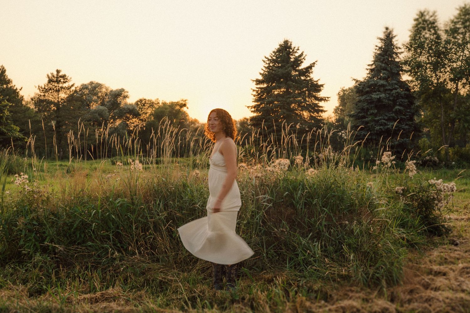 Person in a white dress twirling in a golden field at sunset with evergreen trees in the background.