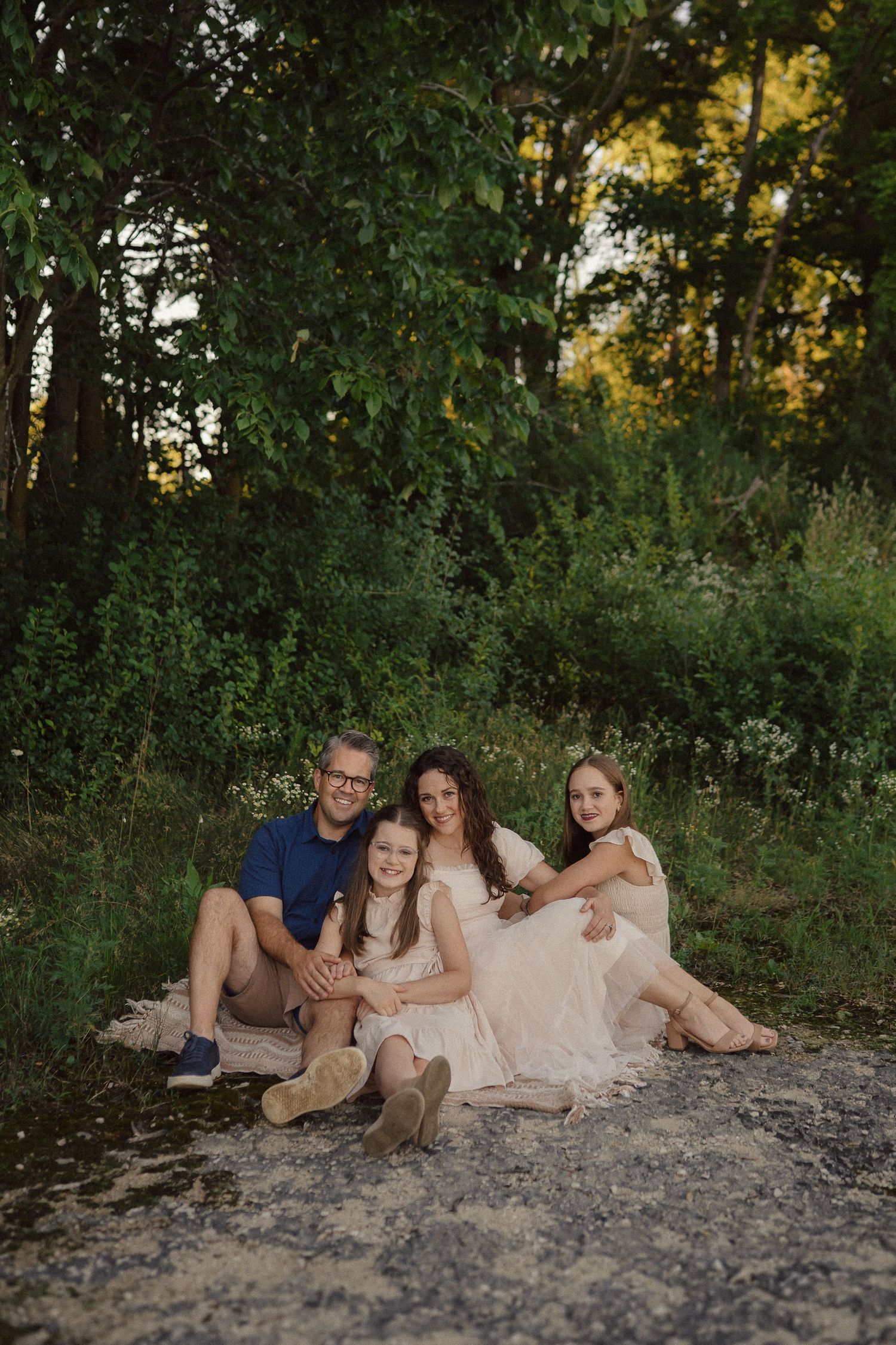 Family relaxes together on rocky ground against lush green forest backdrop.