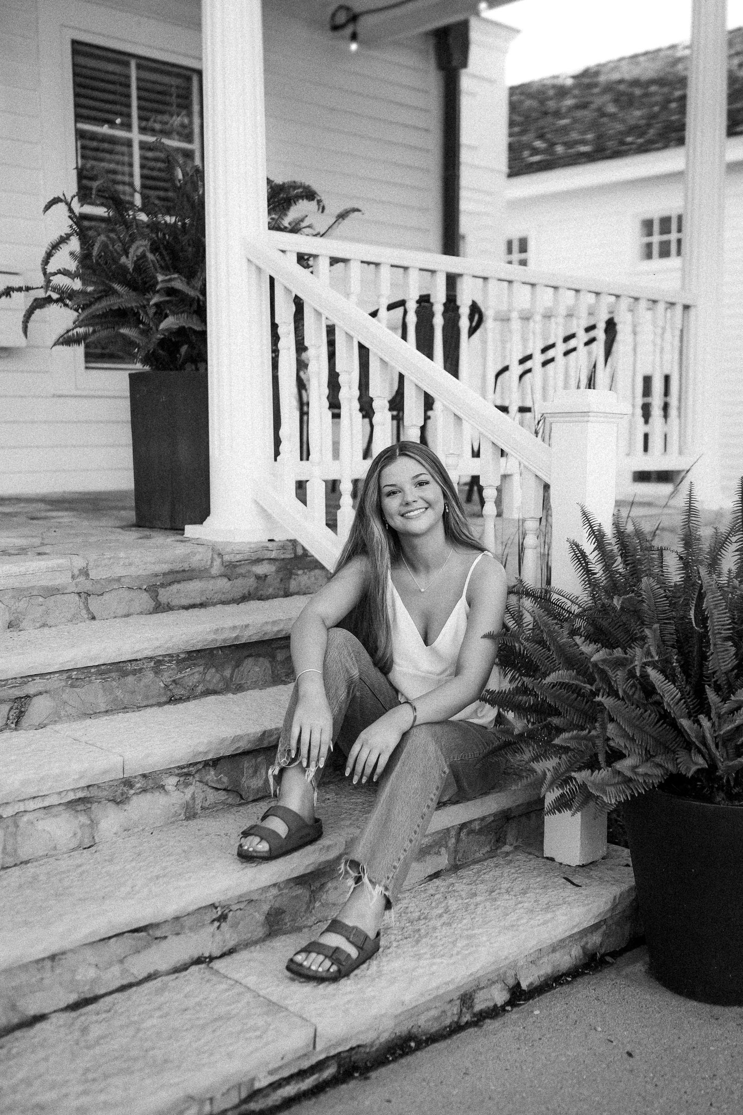 A black and white portrait of a woman sitting on stone steps with potted ferns.