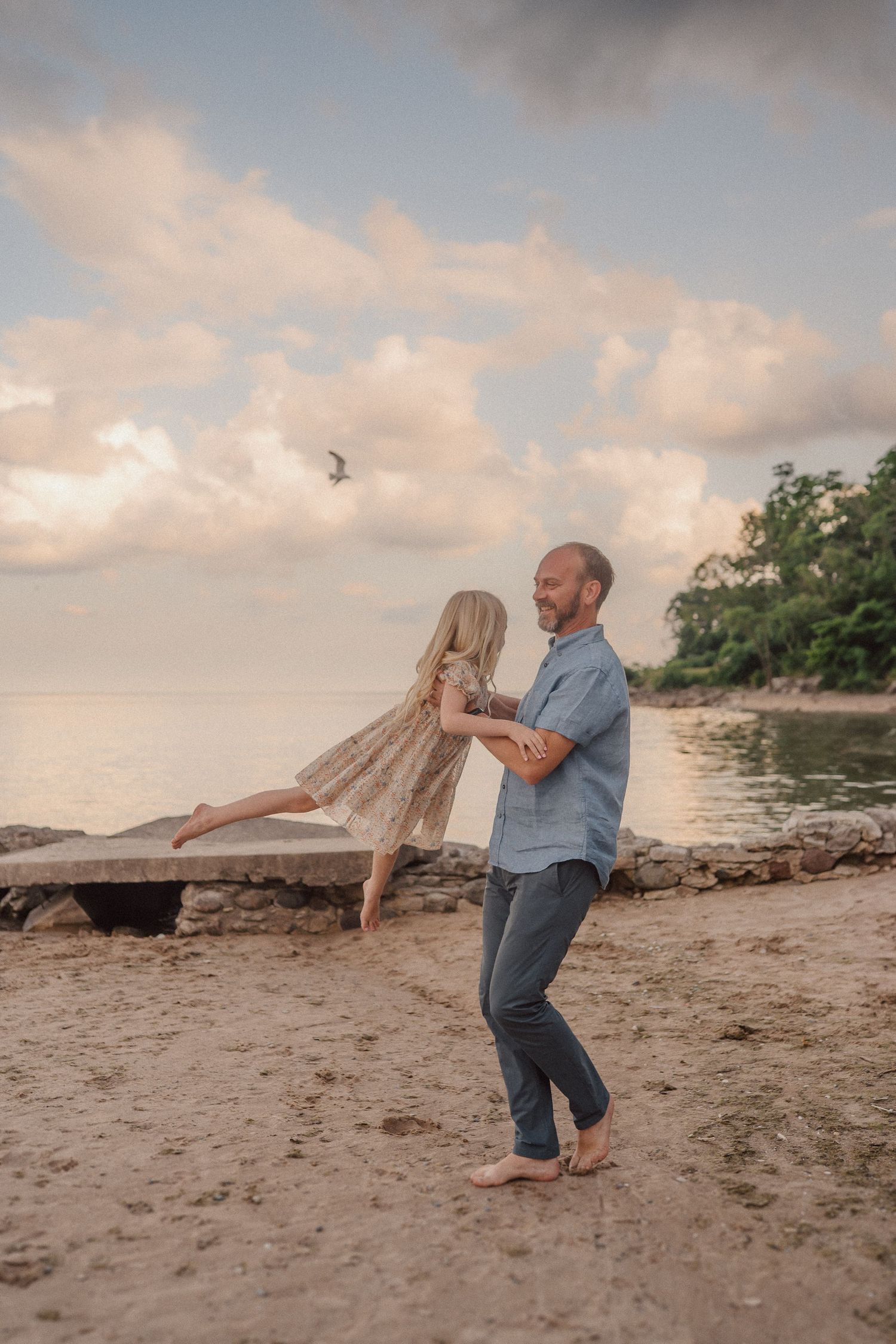 A couple dances together on a beach at sunset with cloudy skies and tropical foliage in the background.