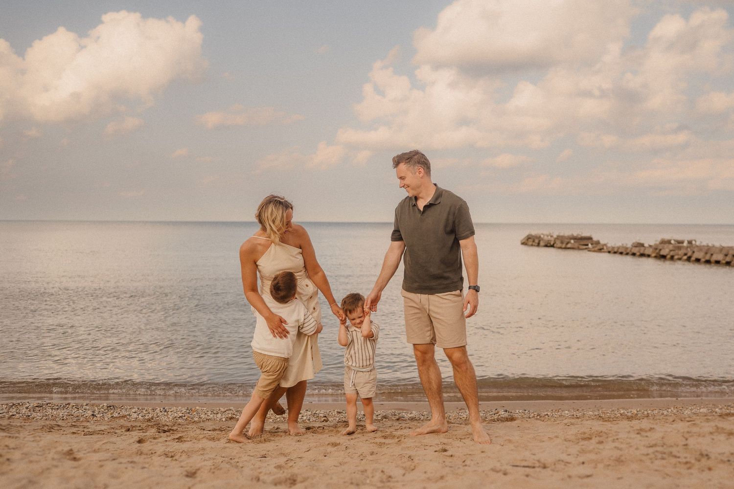 Family walking on scenic beach with jetty during sunset while children play in sand and ocean waves.