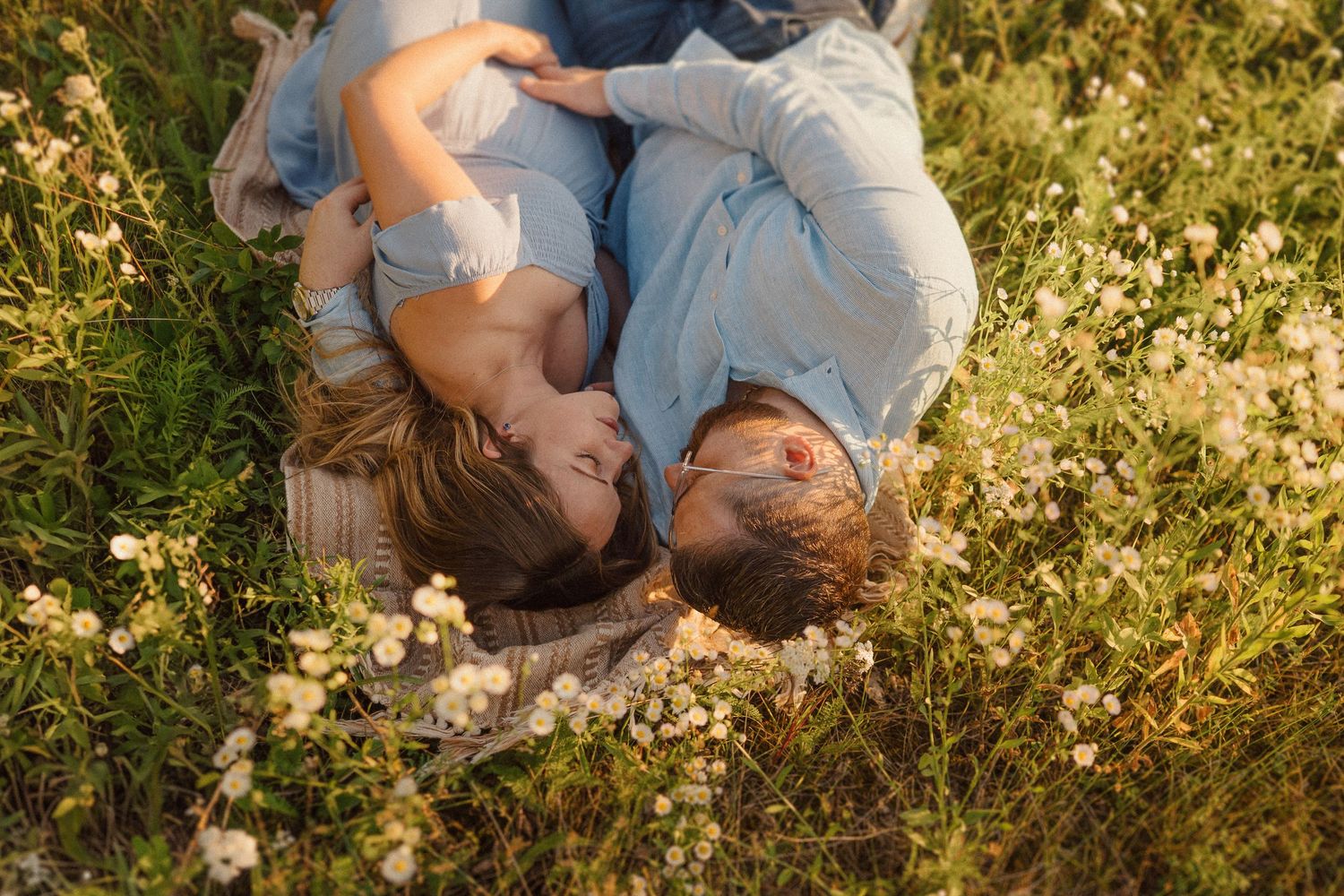 Two people lying in grass with white wildflowers during golden hour in matching light blue outfits.