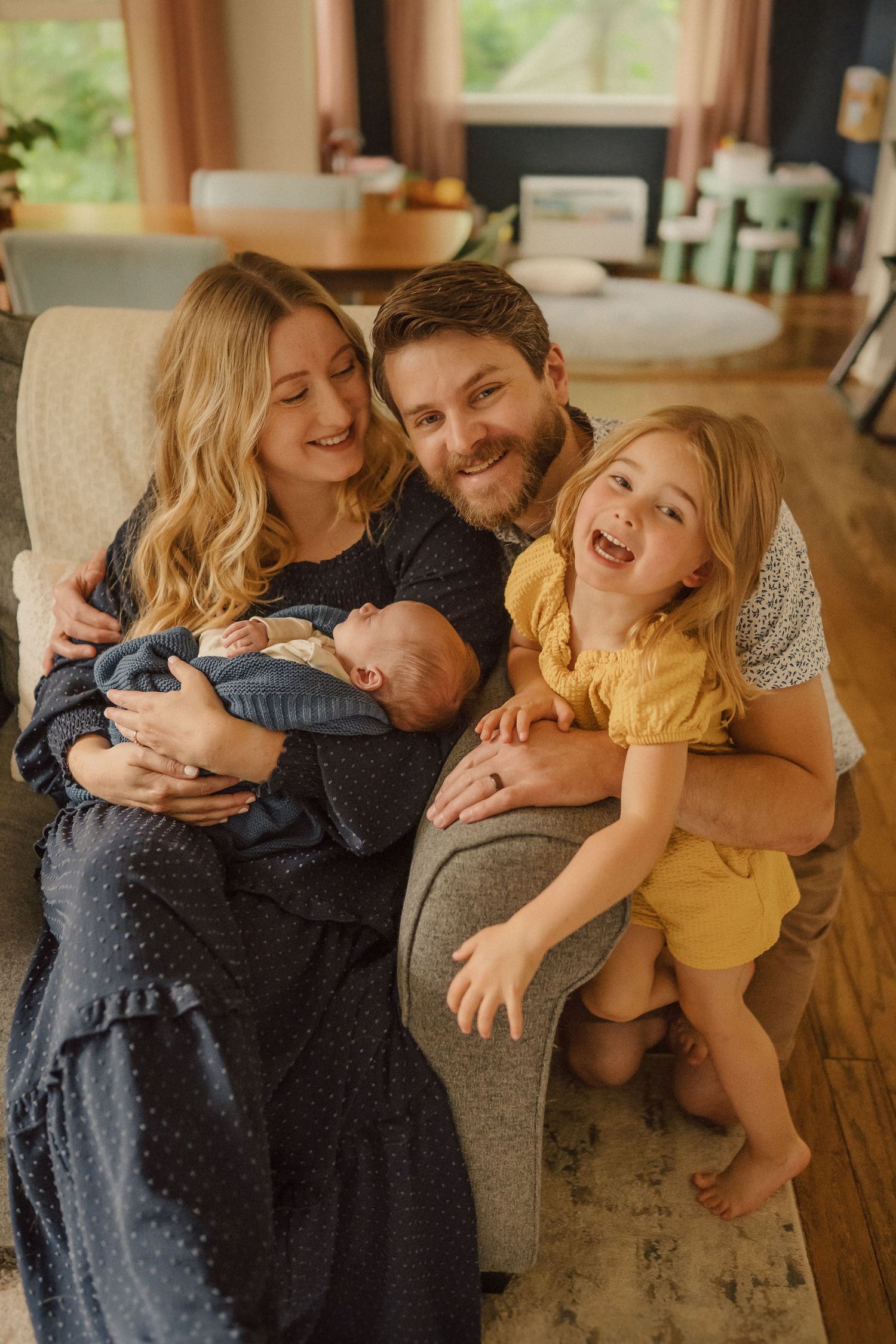 A candid family portrait showing pure joy as they cuddle together with their newborn on a comfortable grey sofa.