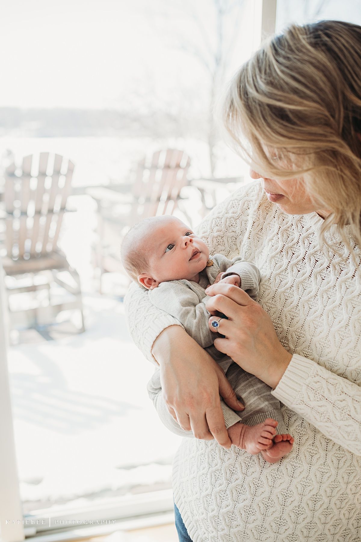 Parent in white sweater gazes lovingly at newborn baby by window with rocking chairs.