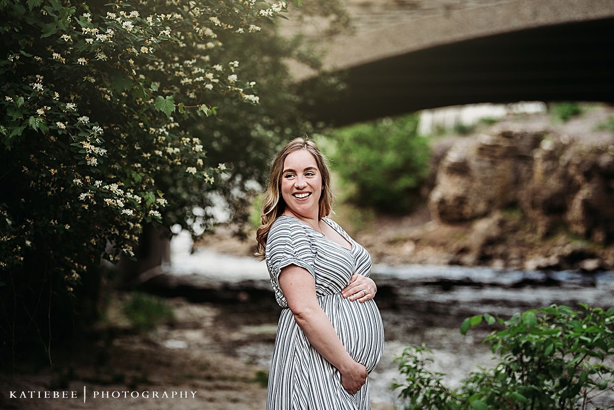 A joyful maternity portrait by a stream with a stone bridge in the background.