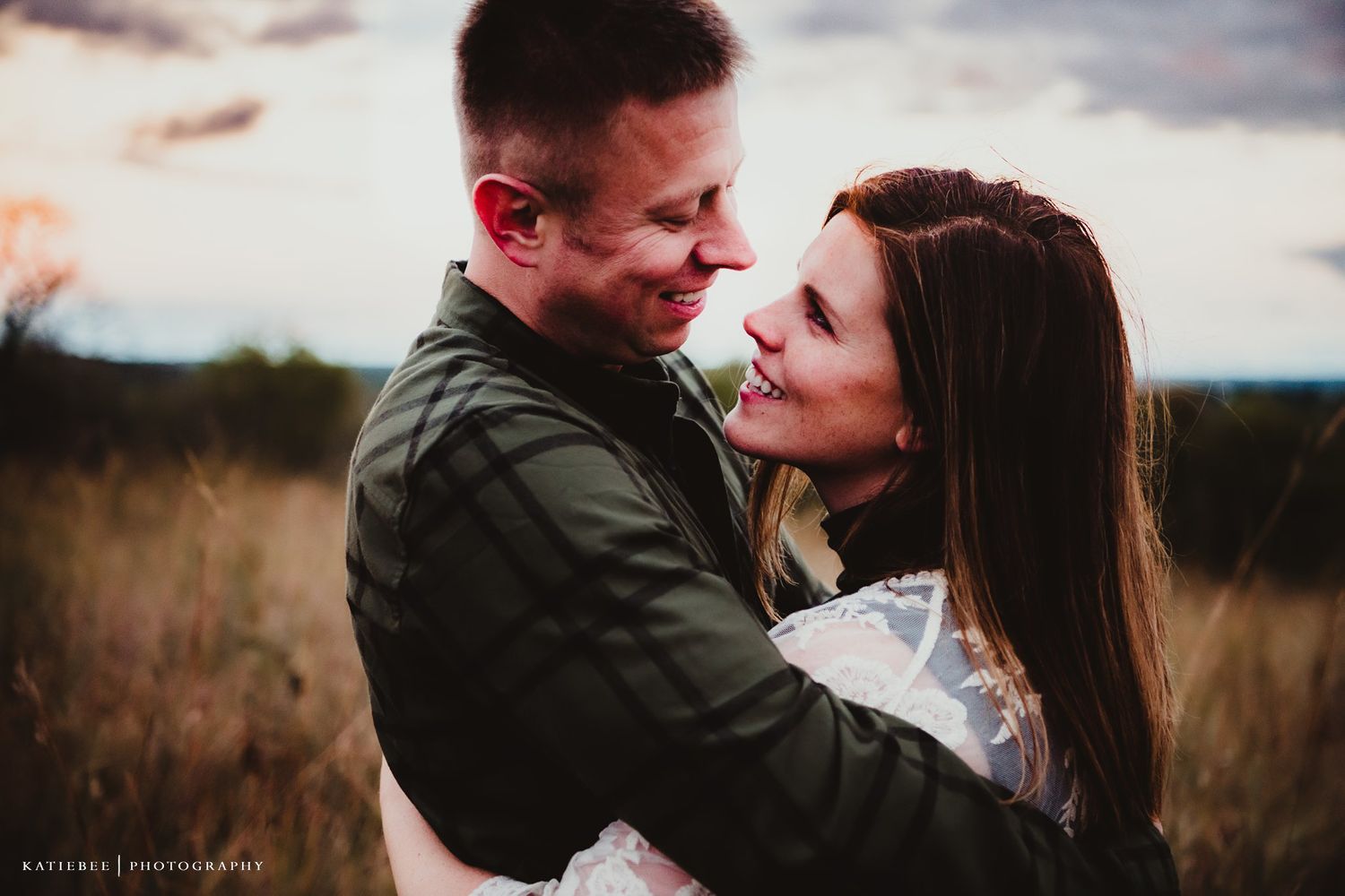 A romantic couple embraces while sharing a laugh in a golden field at dusk.