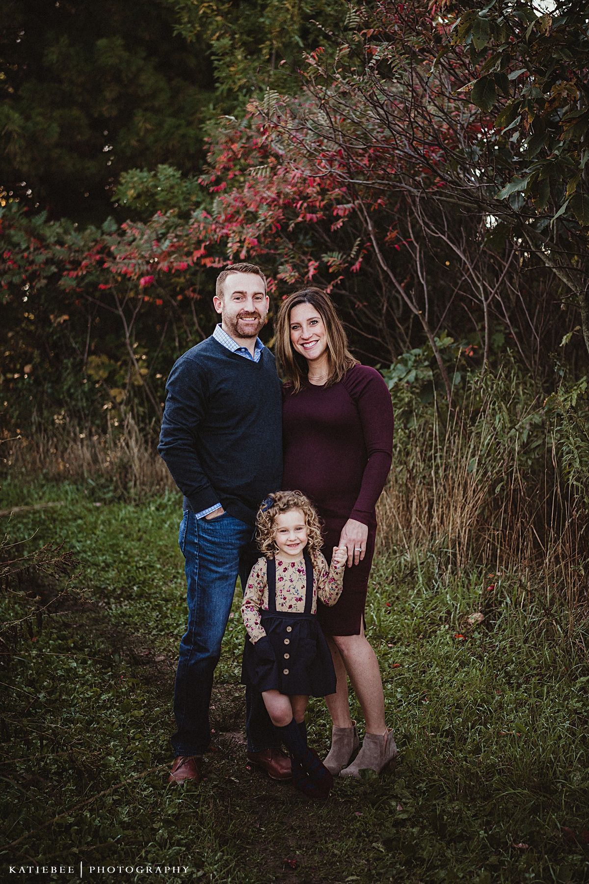 A family of three poses together under red autumn leaves wearing coordinated navy and burgundy outfits.