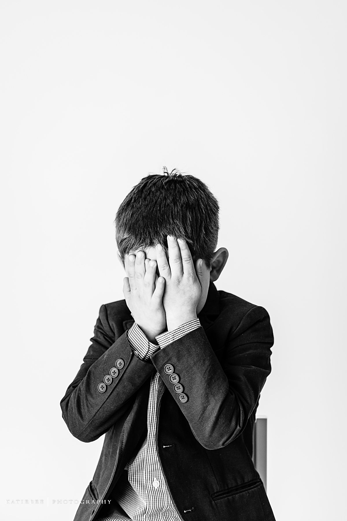 Monochrome portrait of child in blazer covering face with both hands.