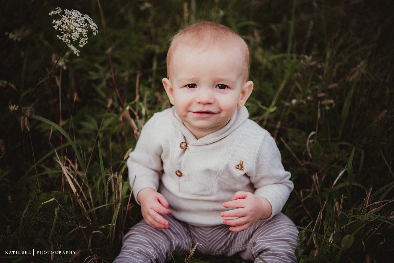 Baby in cream sweater sits in grassy field smiling at camera.
