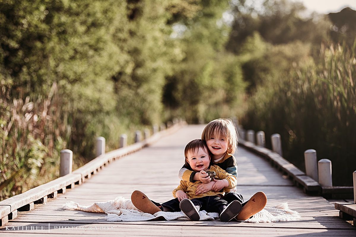 Two children sit close together on wooden boardwalk during sunset photo session.