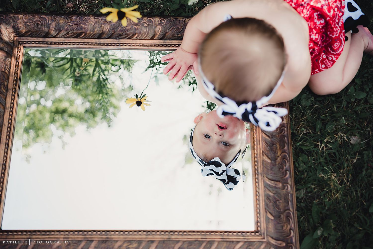 Baby outfit reflected in vintage mirror frame placed on grass outdoors.