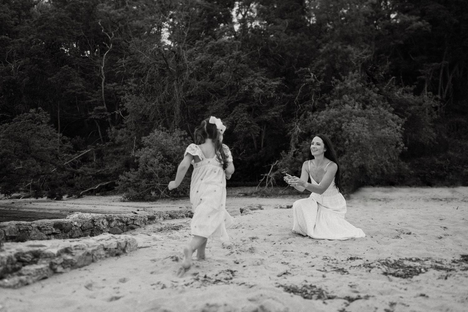 People in flowing dresses dance joyfully on a beach with forest backdrop.