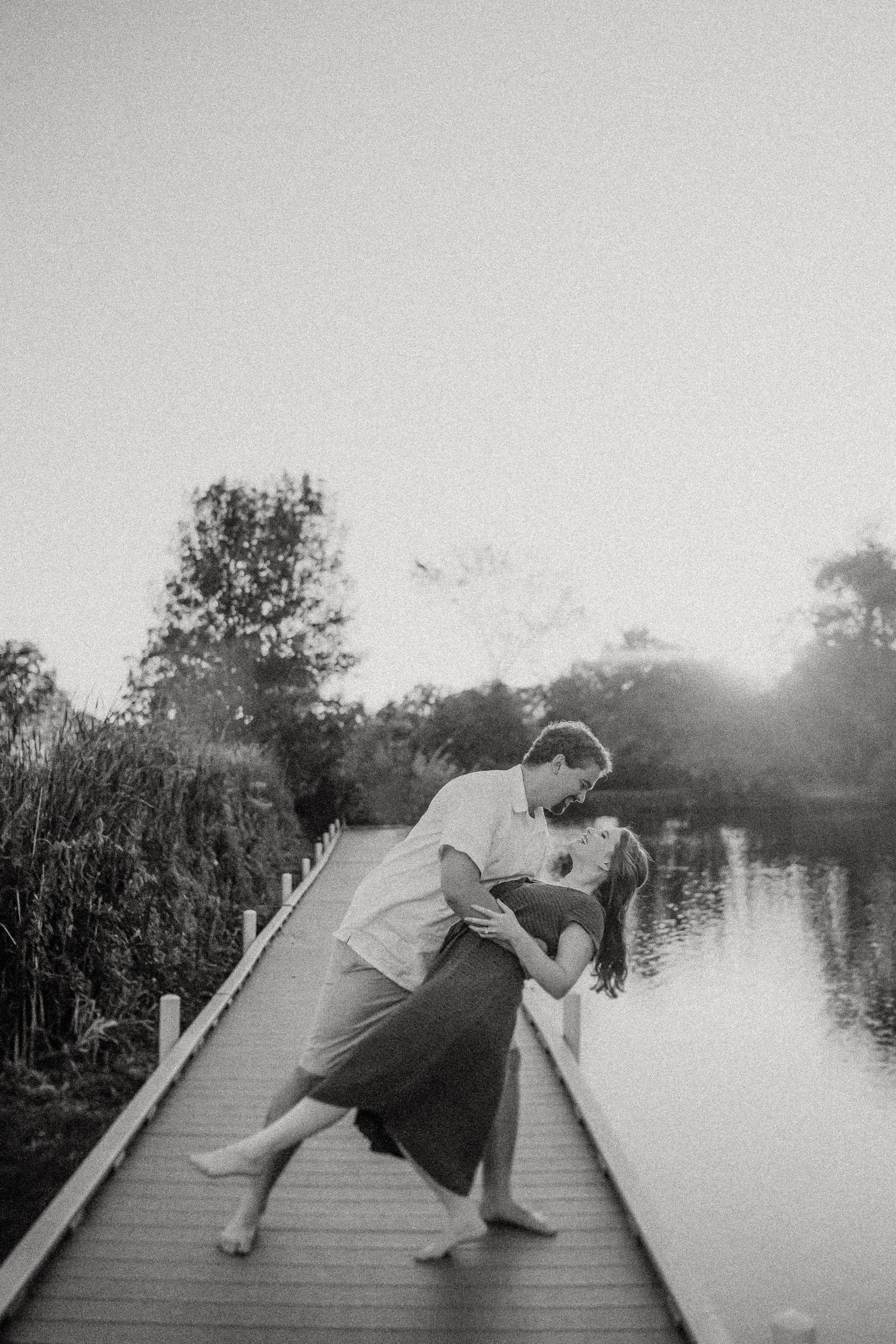 A couple dips while dancing on a wooden boardwalk overlooking water at sunset.