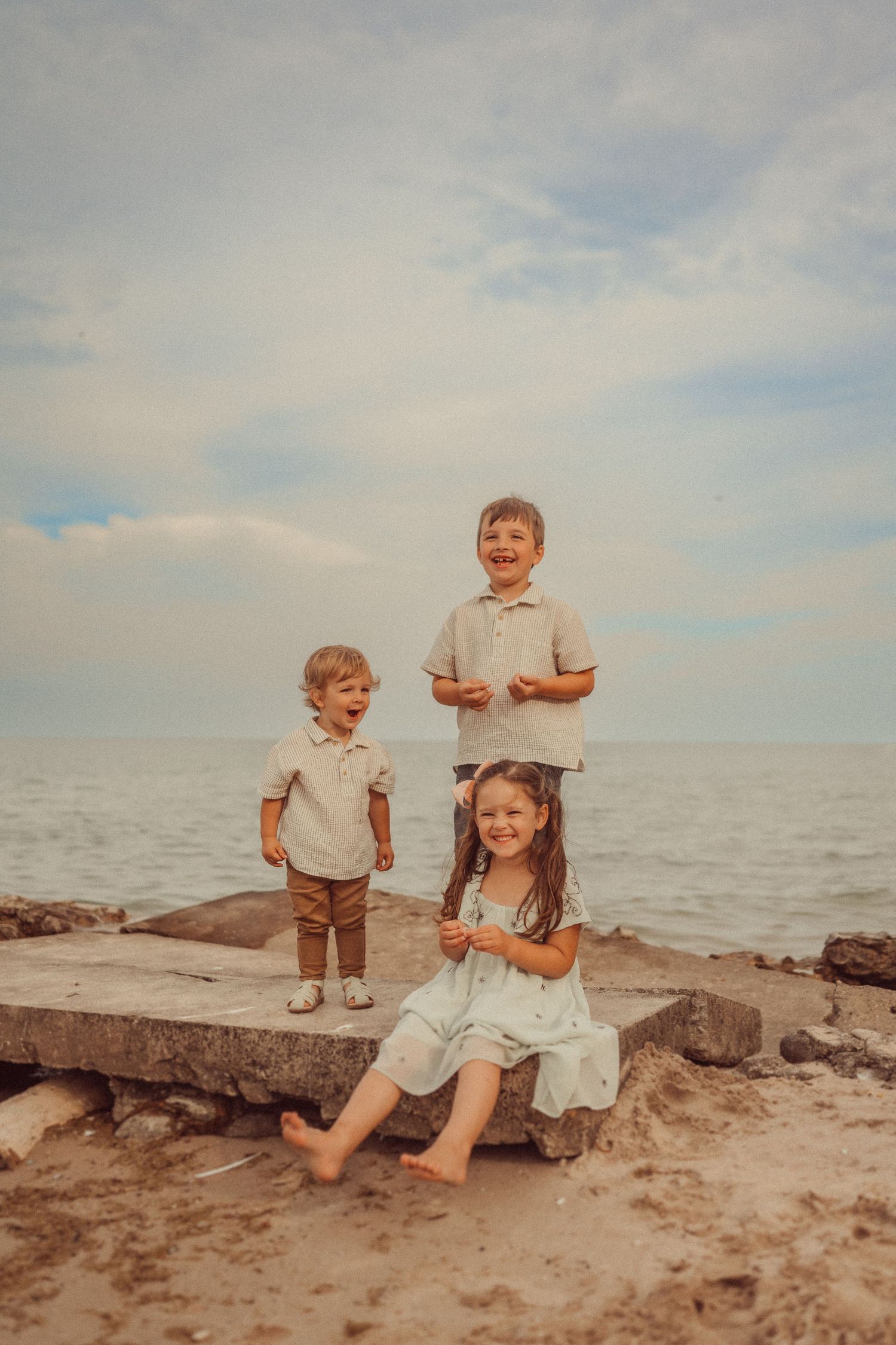 Three children pose on concrete blocks at a rocky beach during sunset.