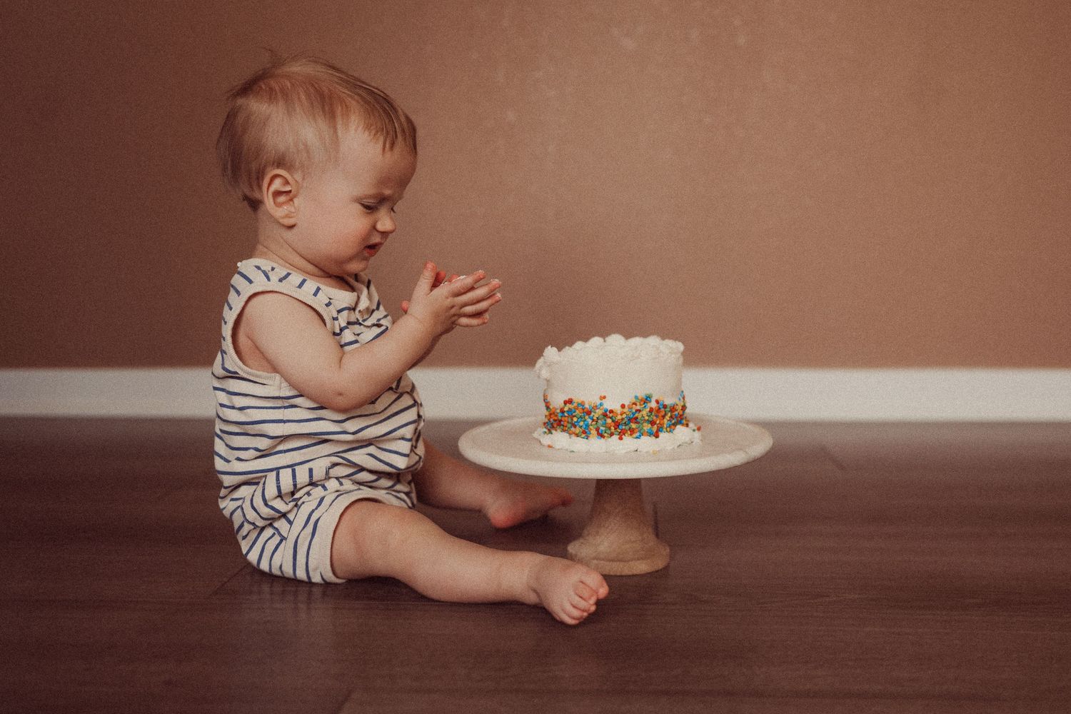 Small child in striped outfit sitting on floor looking at white birthday cake.