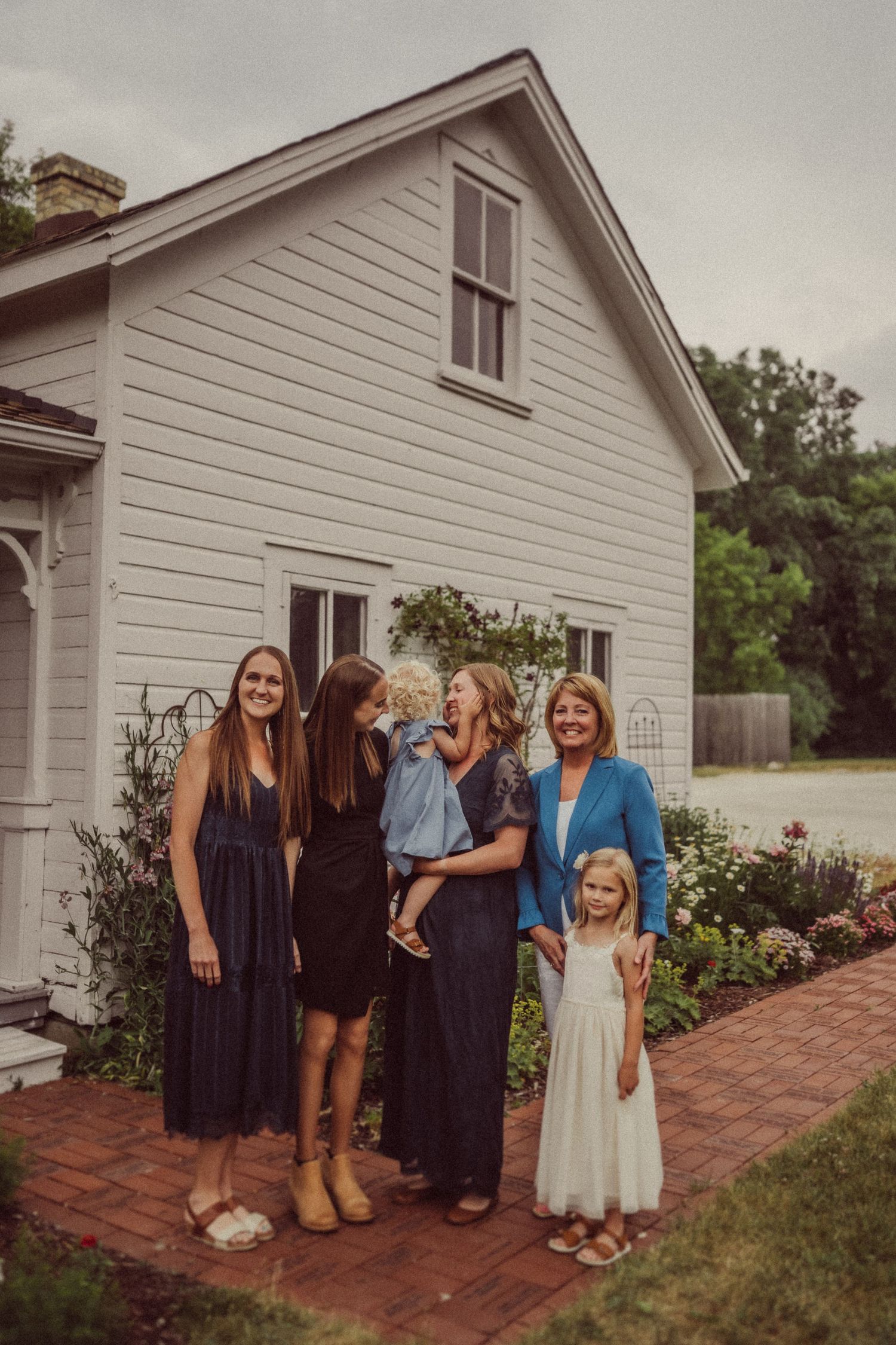 A group stands together in front of a white farmhouse on a brick walkway on an overcast day.