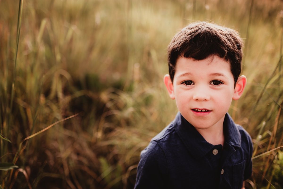 Close-up portrait of a child in a navy polo shirt smiling among tall grass.