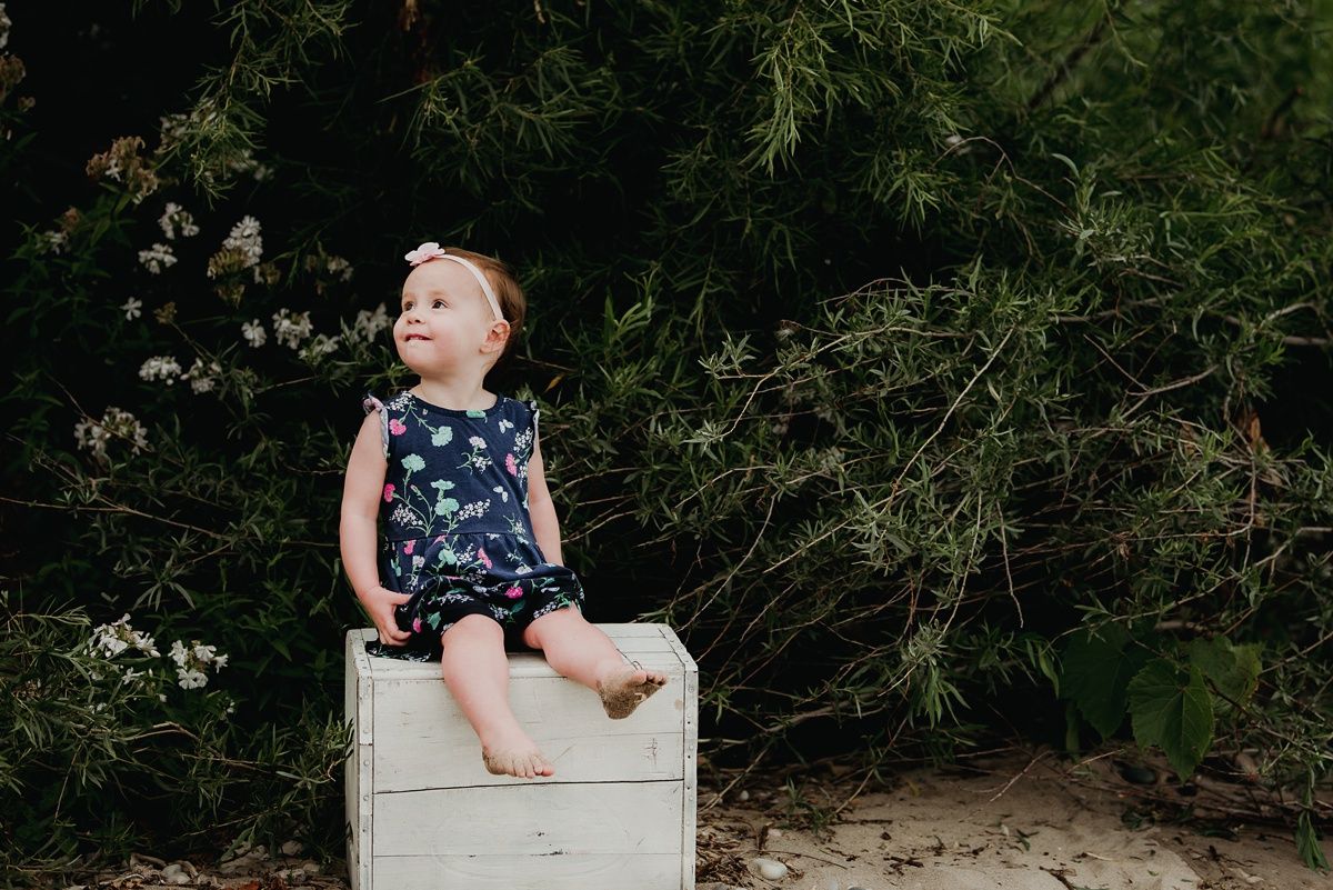 A carefree child in a navy floral dress sits barefoot on a white box outdoors.